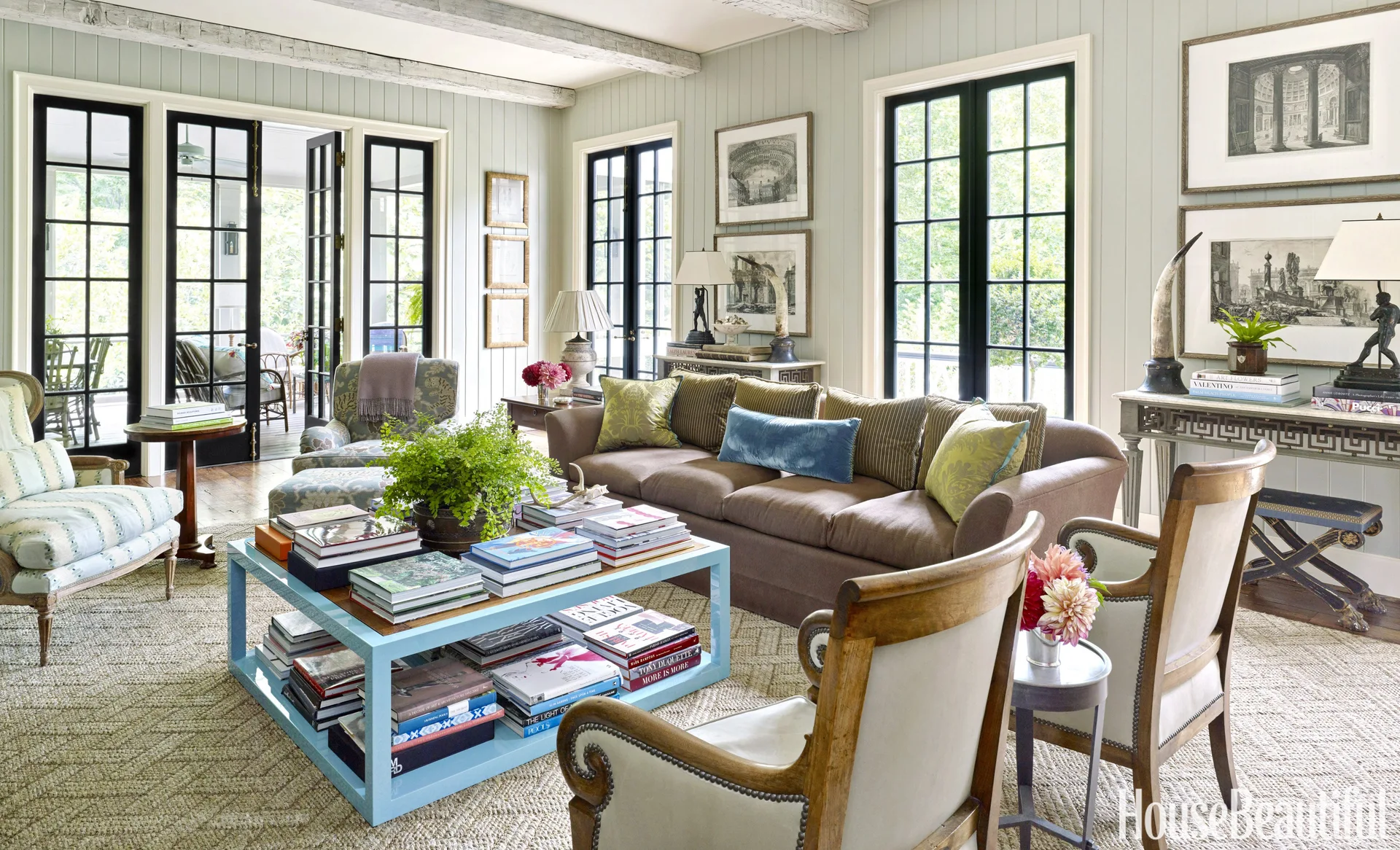 A bright living room featuring sage green vertical wood paneling, a brown sofa, and a light blue tiered coffee table covered with stacks of books.