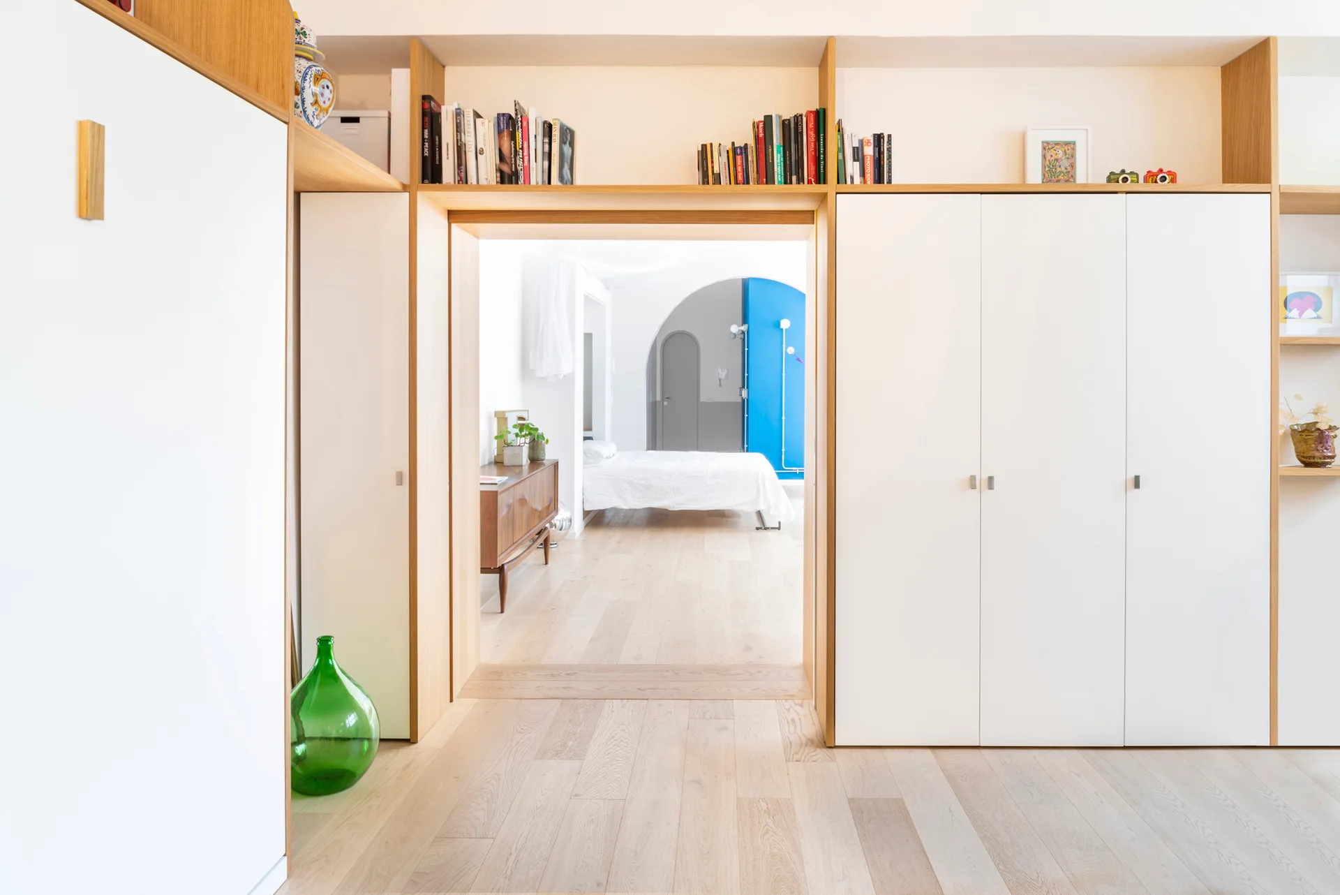 White built-in cabinetry with light wood shelving surrounds a doorway looking into a bright bedroom with light wood floors.