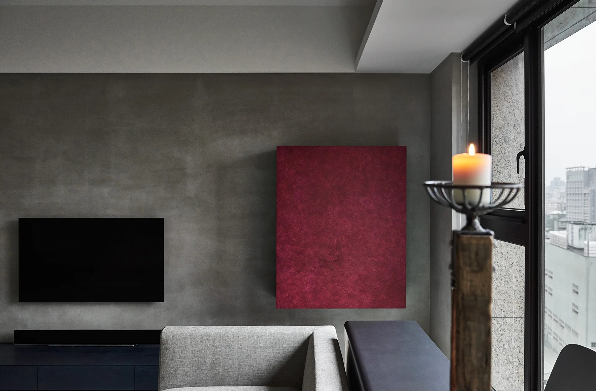 A modern living room featuring a textured grey concrete wall, a wall-mounted TV, a vibrant red square canvas, and a grey sofa.