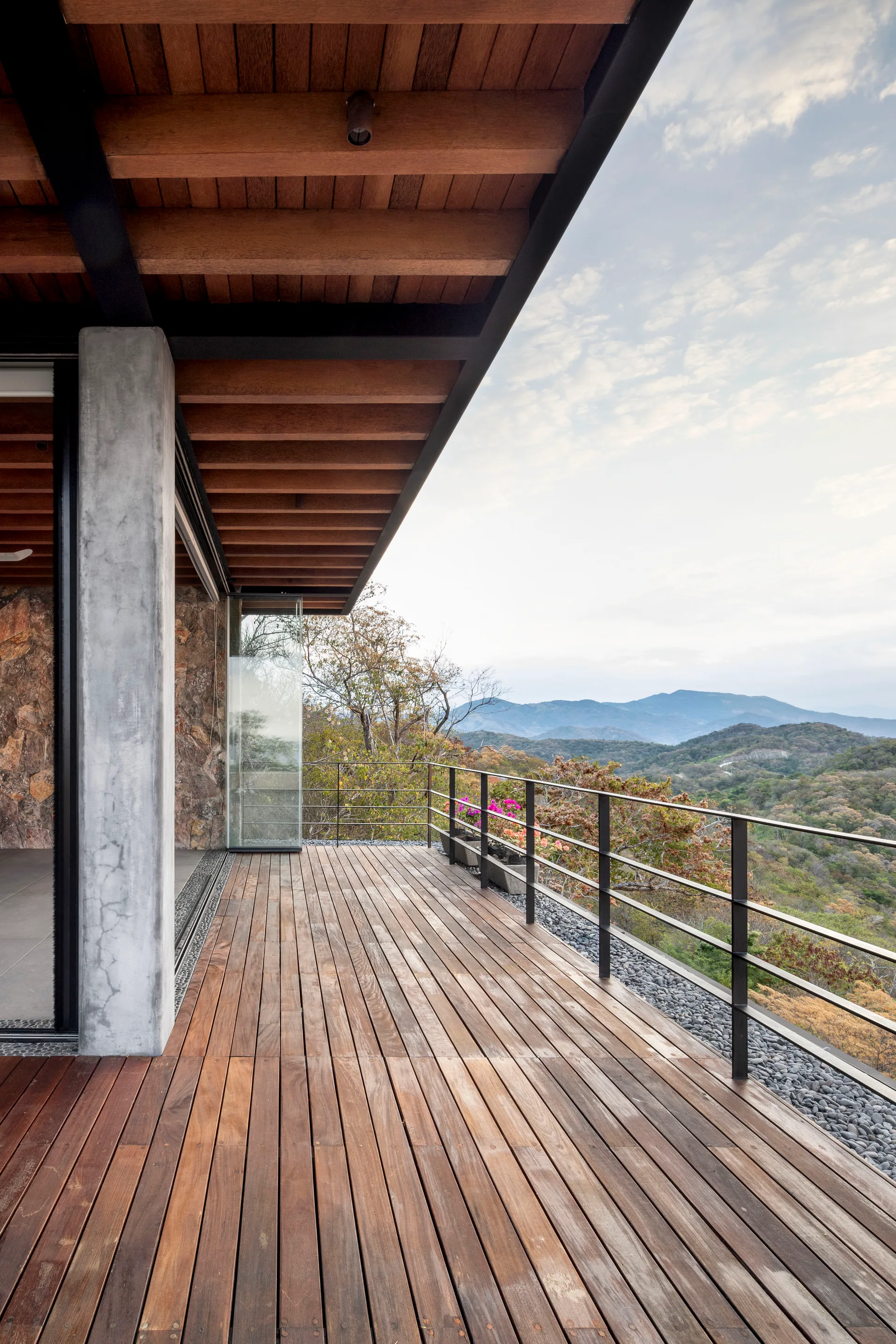 A modern outdoor wooden terrace features a raw concrete pillar, exposed timber ceiling beams, a black metal railing, and a panoramic mountain landscape view.
