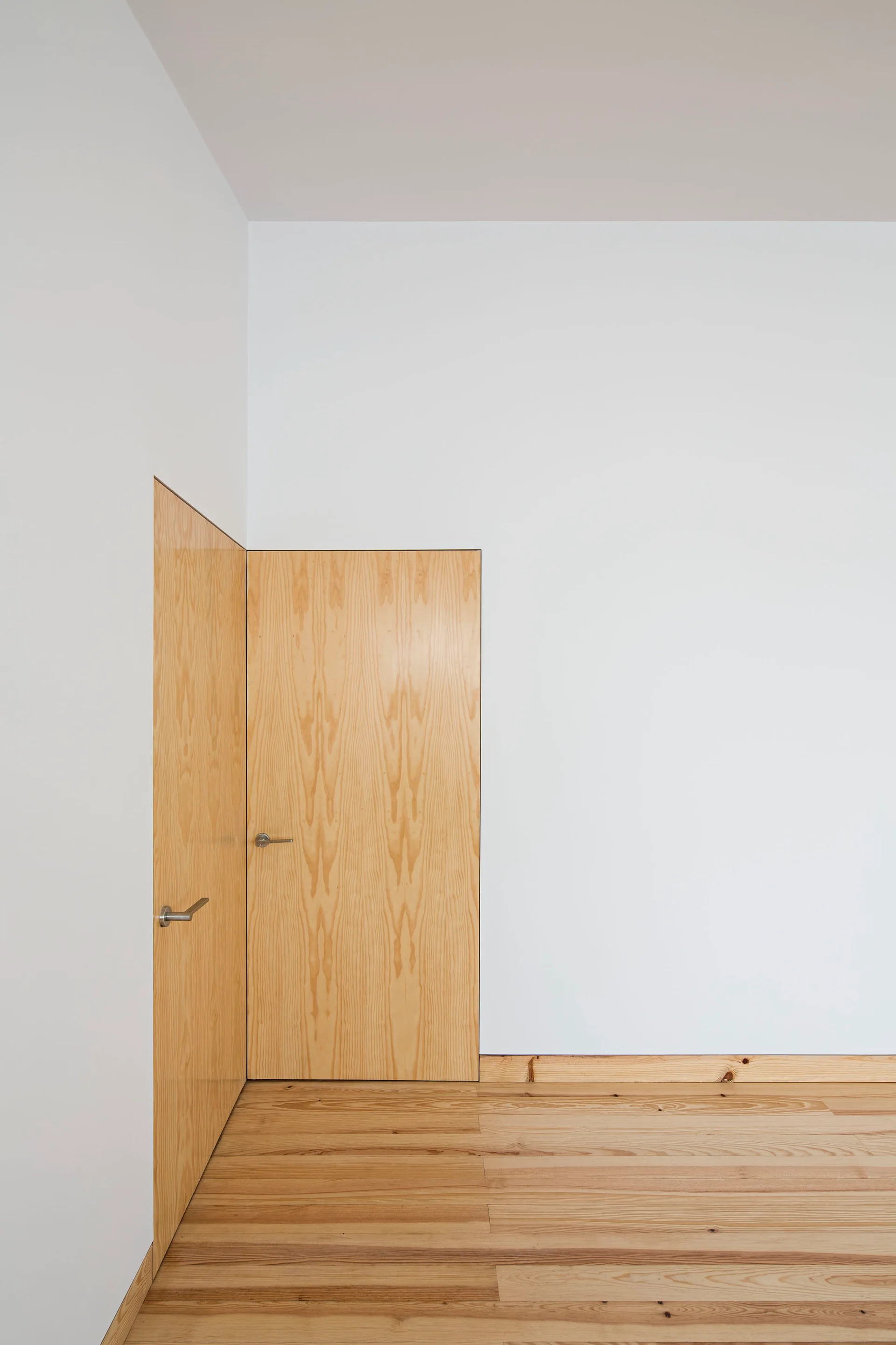 Two flush-mounted light wood veneer doors meet at a corner against plain white walls and matching wood plank flooring.