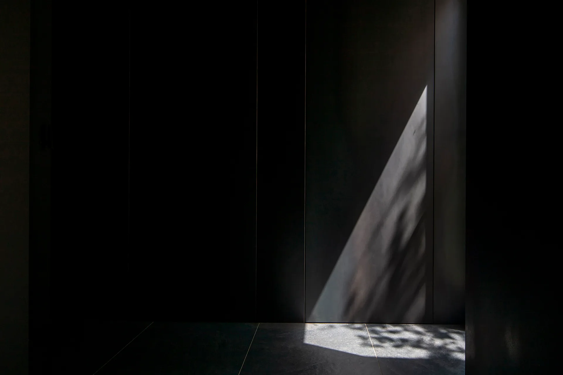 A minimalist interior featuring dark matte vertical wall panels and large grey floor tiles illuminated by a single diagonal beam of sunlight.