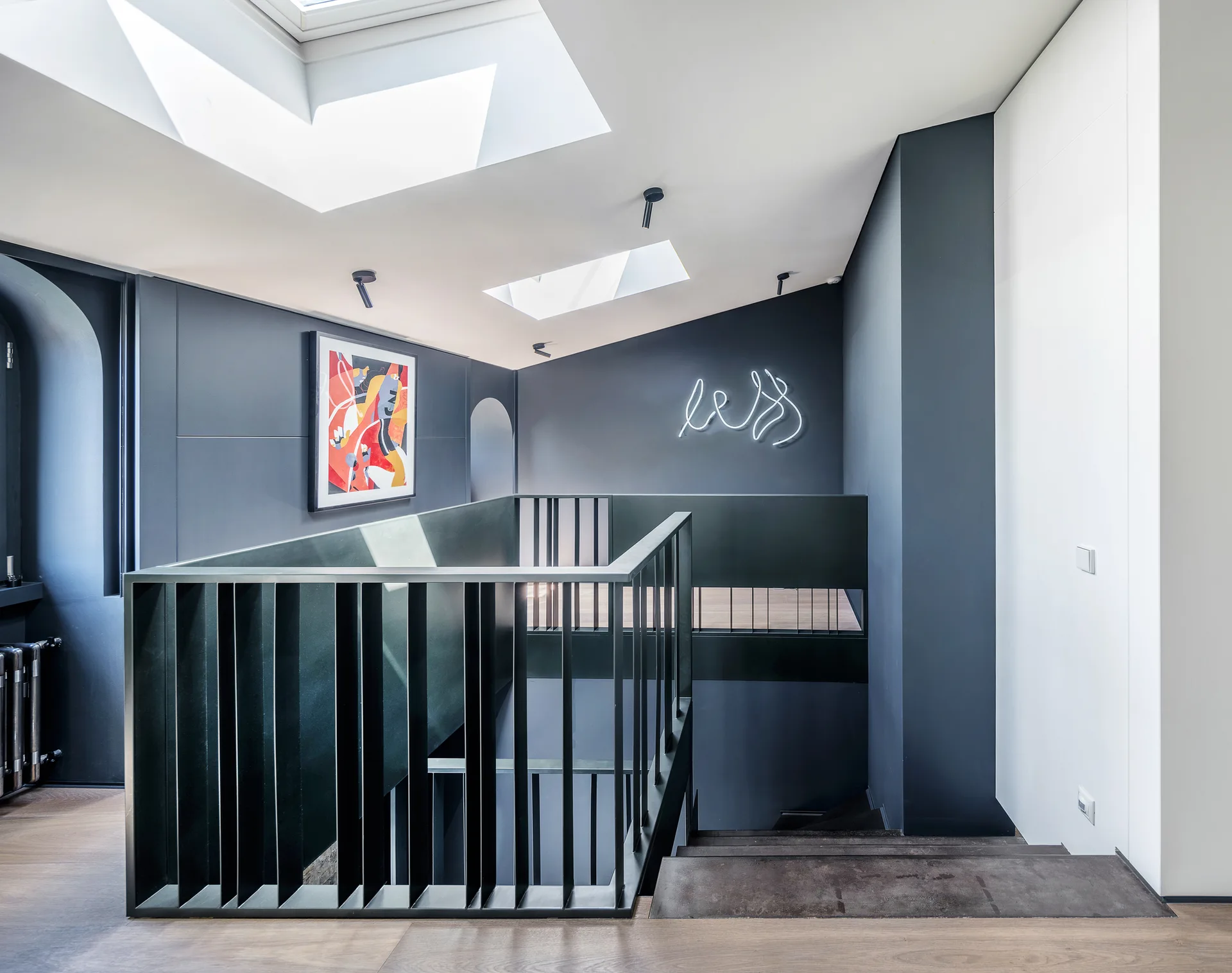 A modern staircase landing with dark gray walls, a black metal slatted railing, overhead skylights, and a glowing neon sign.