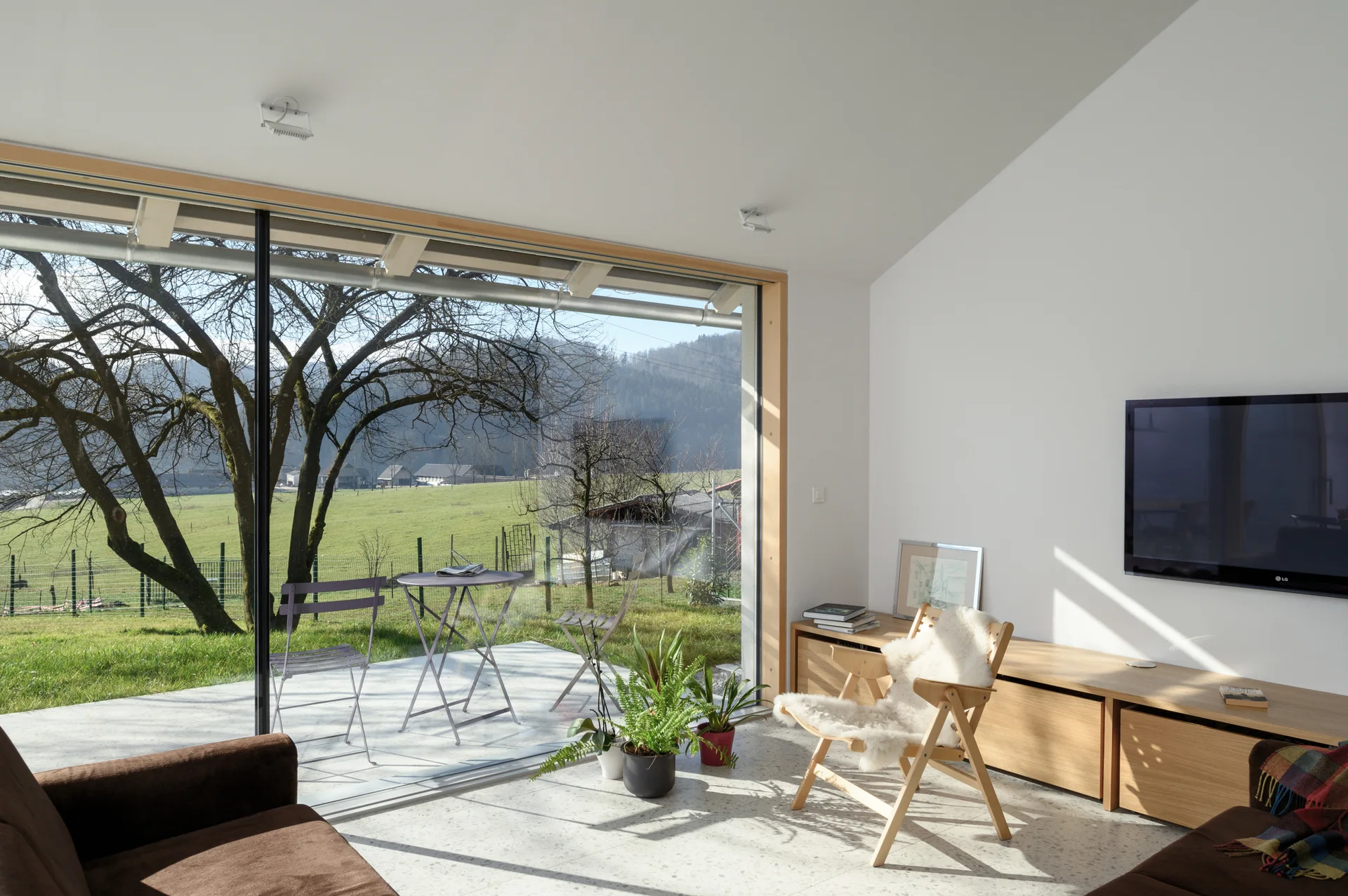 A minimalist living room with a low wood media console, a wooden chair with a sheepskin throw, and a large sliding glass door opening to a green terrace.