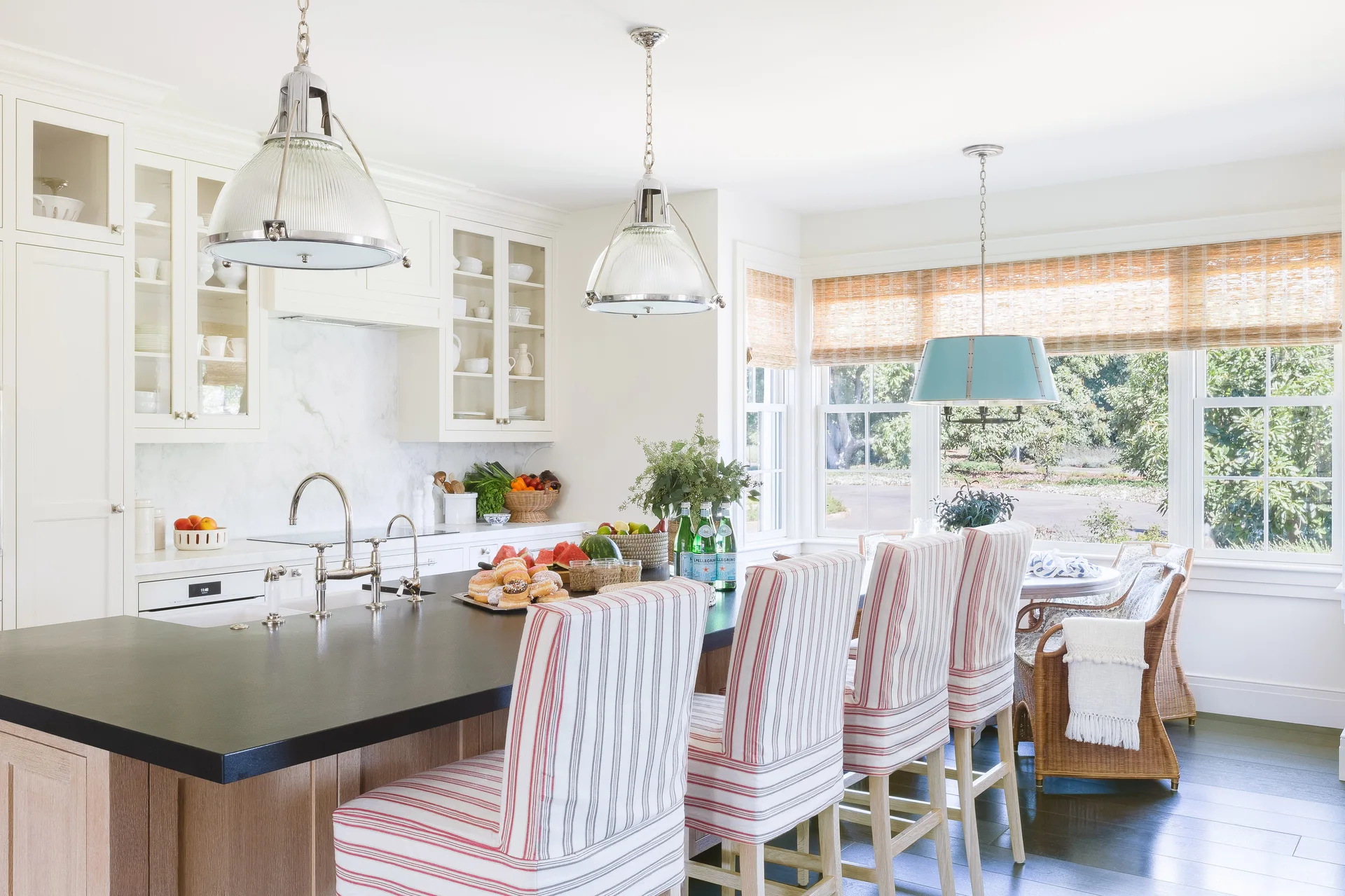 A white coastal kitchen featuring a large black countertop island with striped bar stools and glass dome pendant lights.