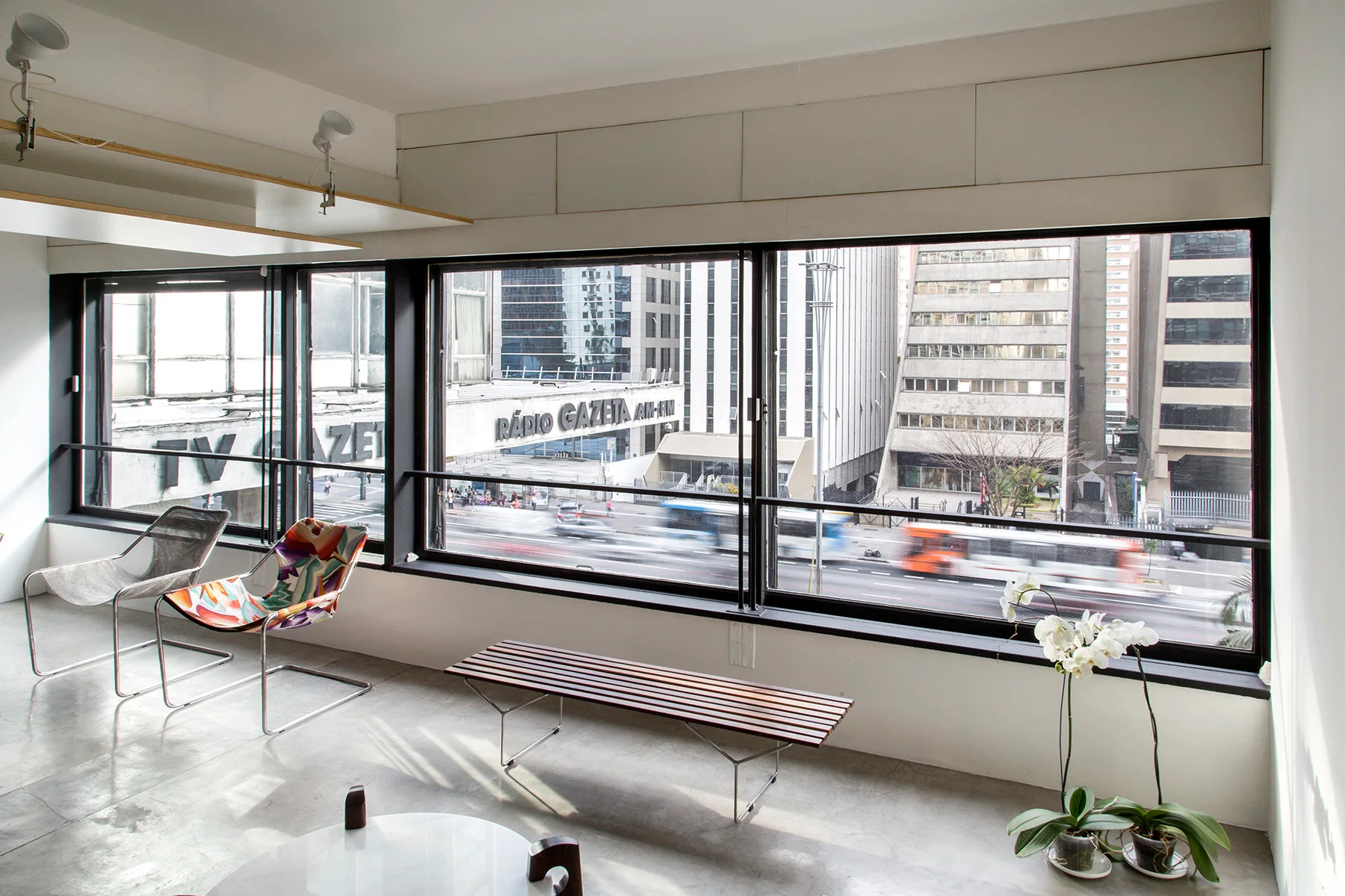 A minimalist living area features concrete flooring, a slatted wood bench, and wireframe chairs in front of large windows overlooking a city street.
