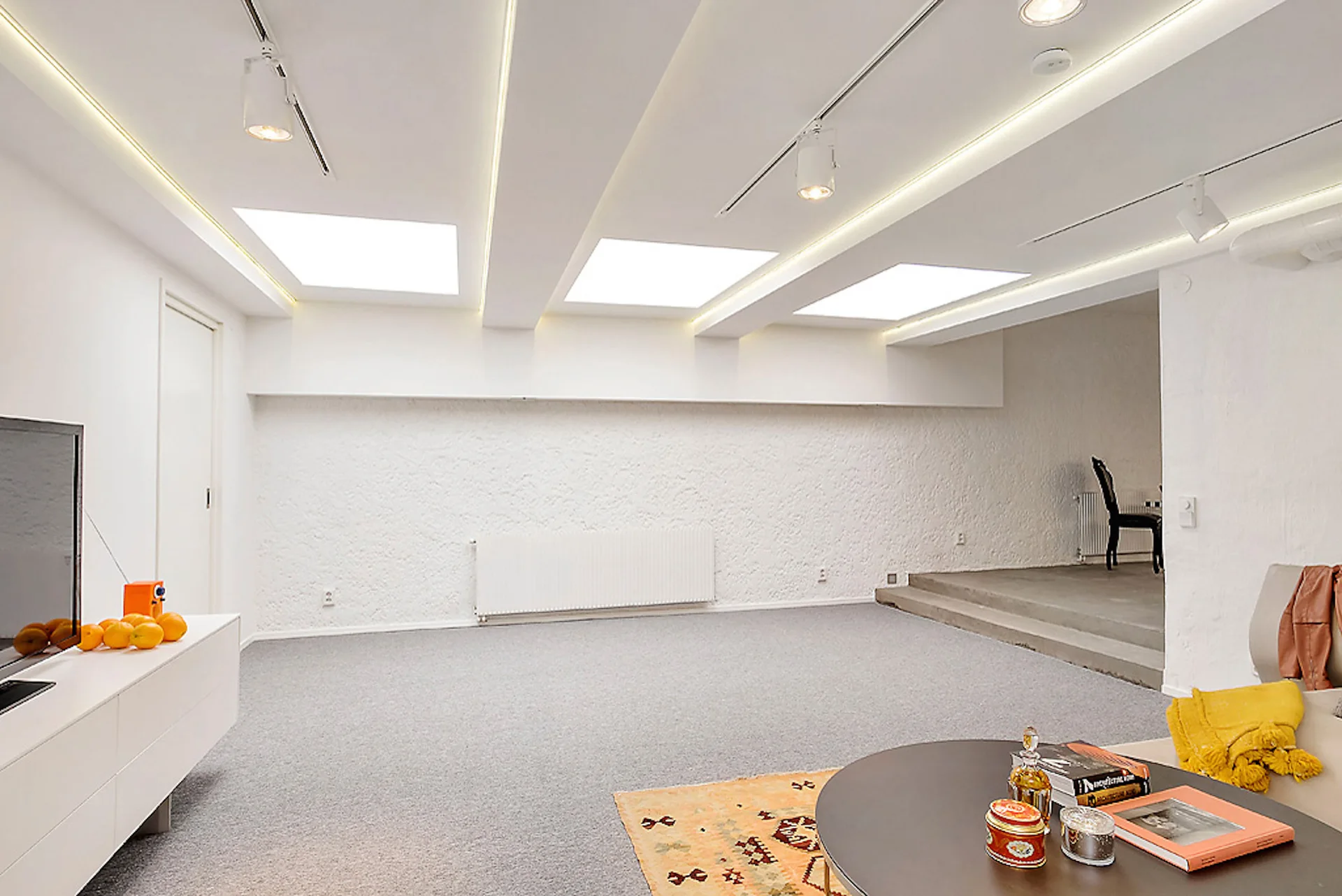 A minimalist split-level living room features three square skylights, track lighting, white textured stucco walls, and grey carpet.