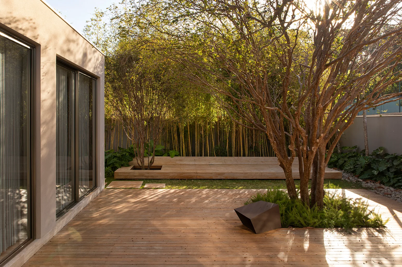 A modern outdoor courtyard featuring a wooden plank deck, mature trees integrated into the landscaping, and a beige building facade with glass doors.