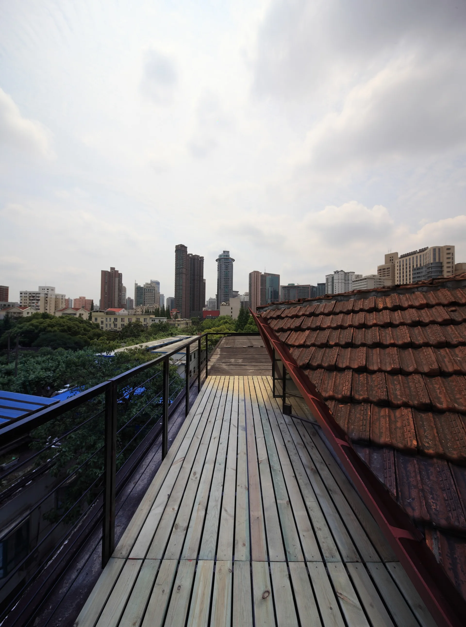 A narrow wooden plank deck with a black metal railing runs alongside a terracotta tiled roof, overlooking a city skyline under a cloudy sky.