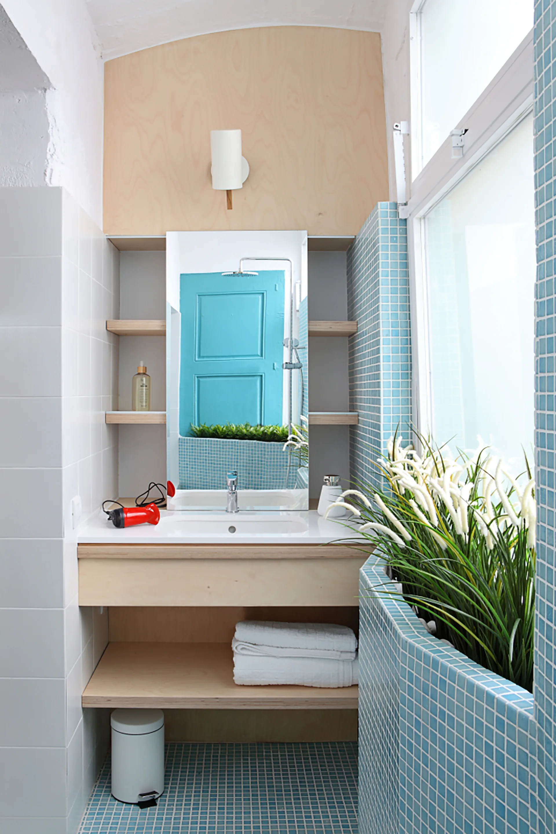 A small modern bathroom featuring a light plywood vanity with open shelves, a tall mirror, and floors and walls covered in blue square mosaic tiles.