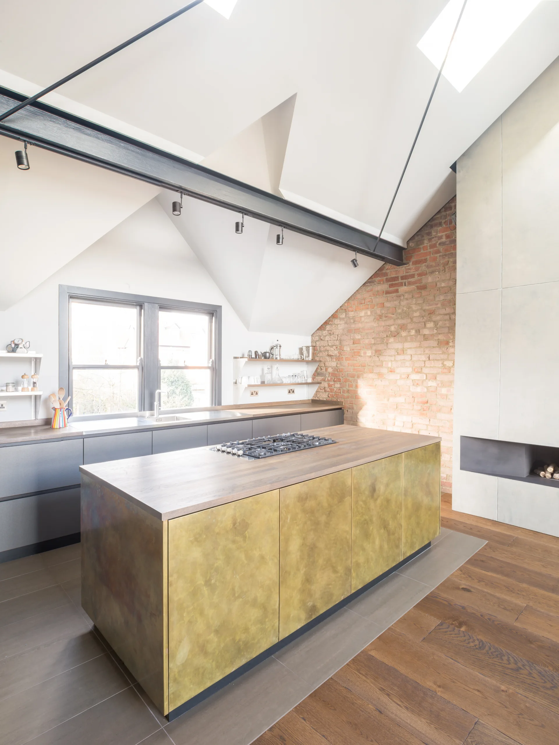 A modern industrial kitchen featuring a brass-finished island, dark grey cabinets, an exposed brick wall, and skylights under a sloped ceiling.