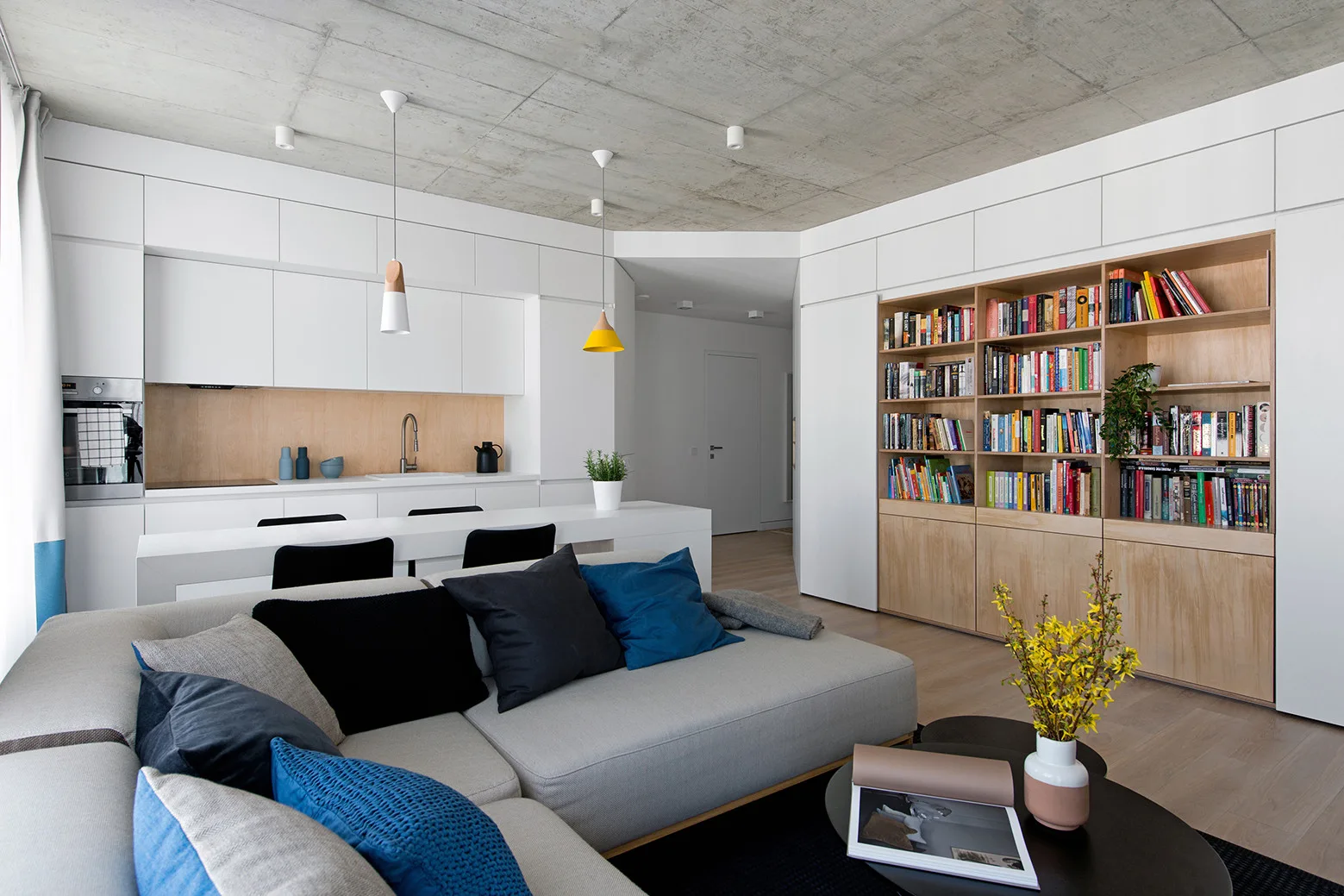 An open-plan living room and kitchen featuring a grey modular sofa, built-in wooden bookshelves, and an exposed concrete ceiling.