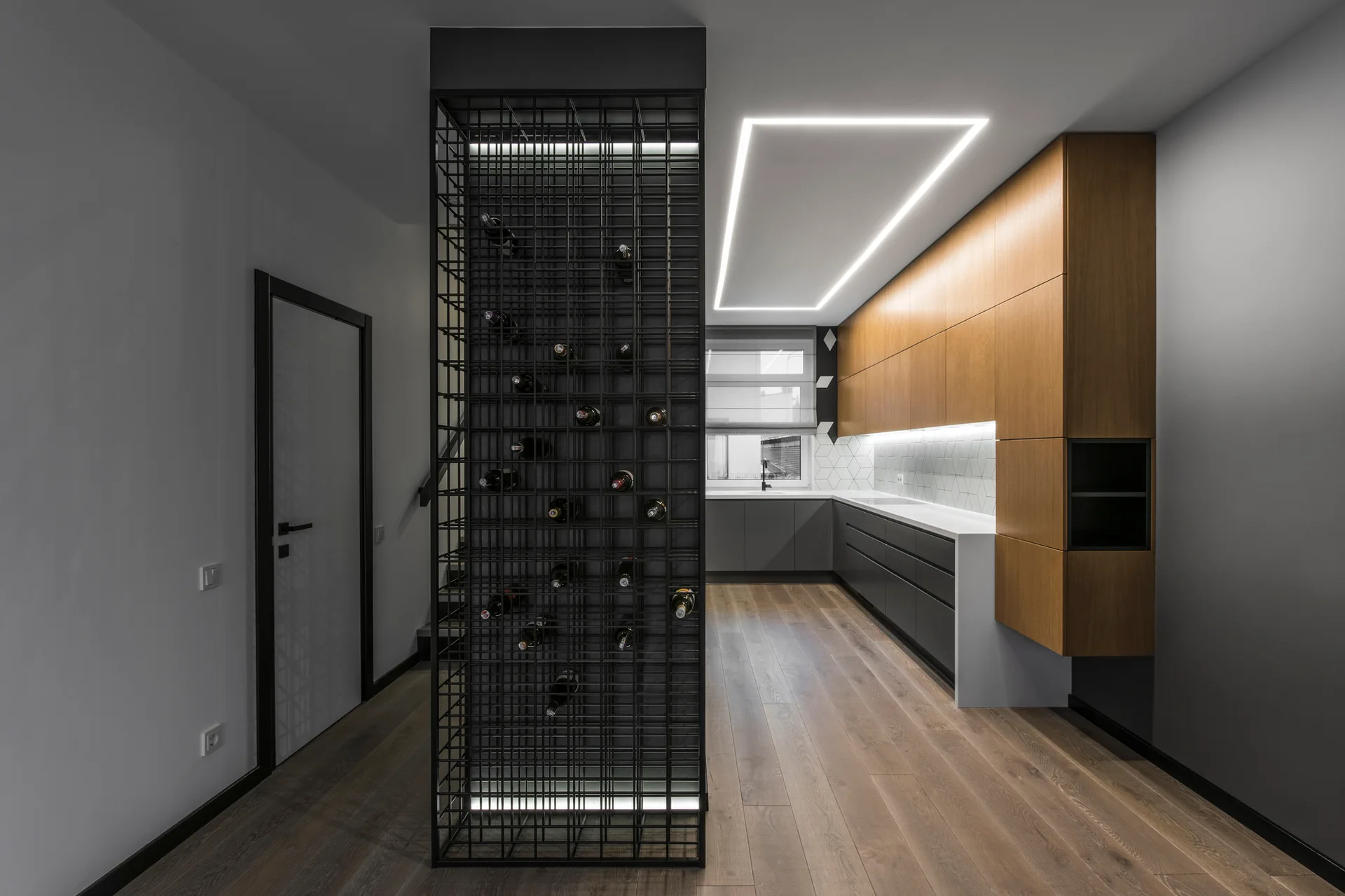 A modern kitchen featuring a black metal grid wine rack partition, wood and charcoal cabinets, and wood flooring under a rectangular ceiling light.