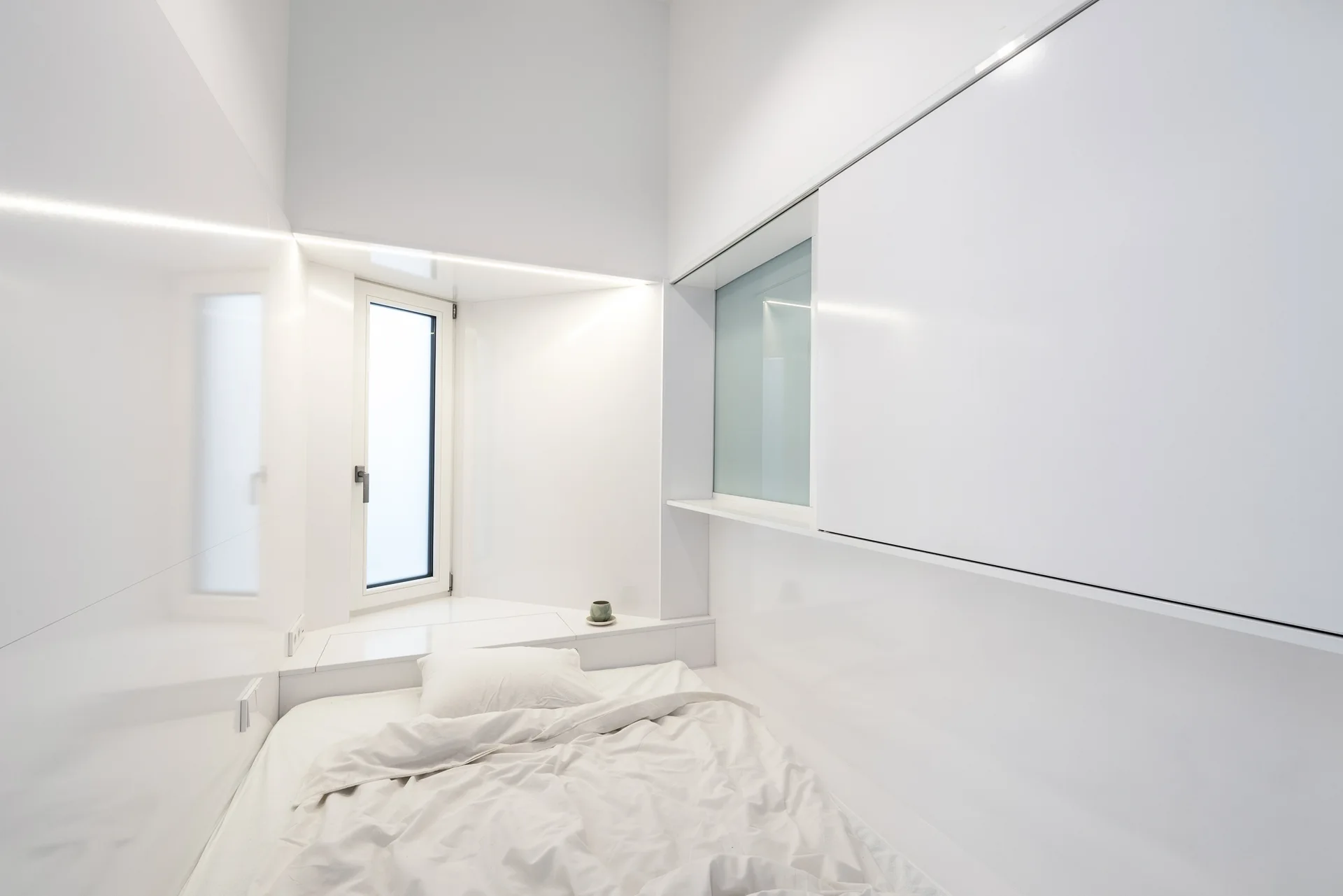 A small minimalist bedroom features a bed with white sheets surrounded by glossy white walls, built-in cabinets, and a window alcove.