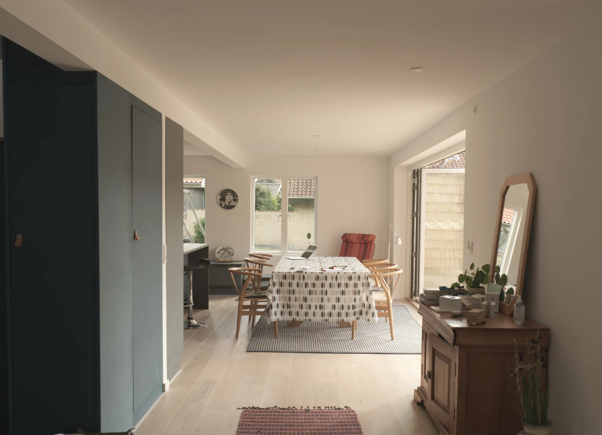 A bright dining room features a wooden table with a patterned tablecloth, surrounded by wooden chairs, a tall blue cabinet, and a wooden sideboard.