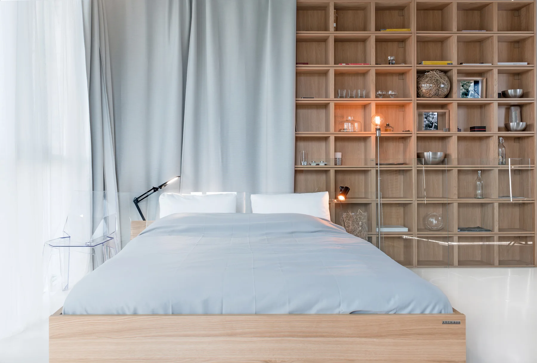 A minimalist bedroom featuring a low light-wood bed with grey bedding, a full-height wooden grid shelving unit, and grey drapery.