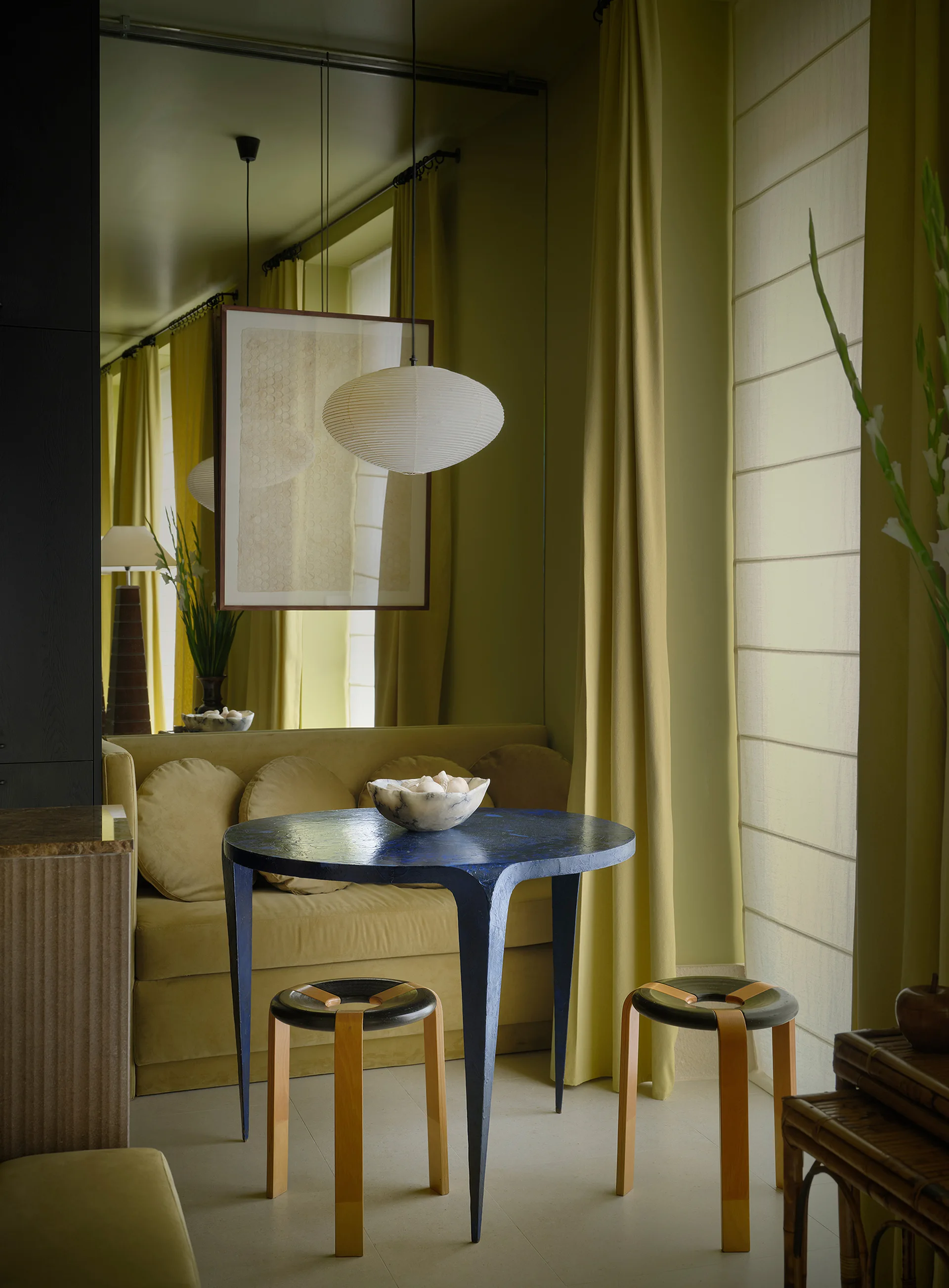 An olive green dining nook features a textured blue round table, two wooden stools, a mirrored wall, and a paper globe pendant light.