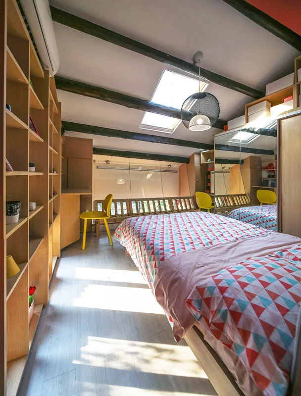 A loft bedroom featuring a bed with colorful geometric bedding, built-in wooden bookshelves, and skylights set between dark ceiling beams.