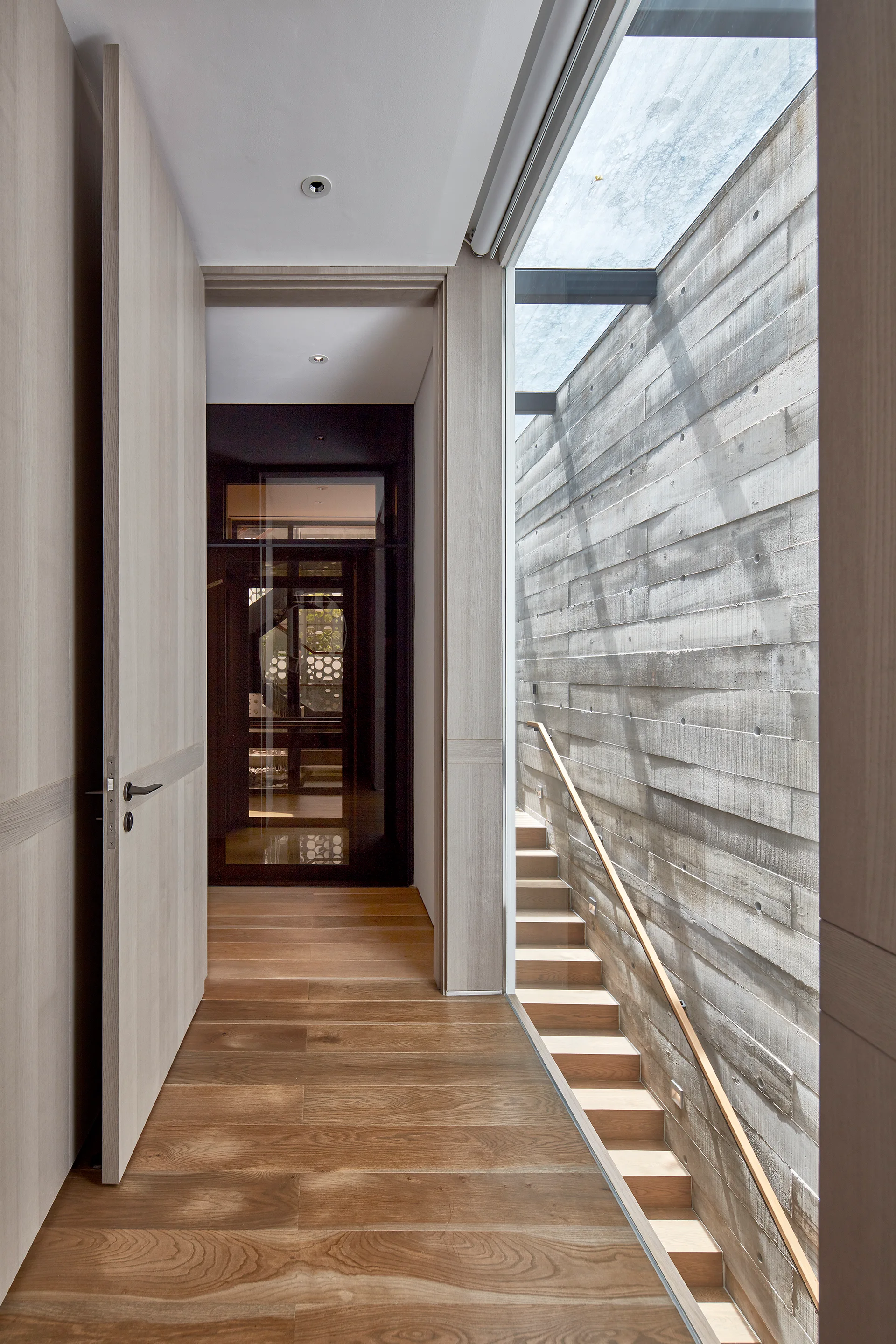 A modern hallway with wood floors features an open doorway leading to a staircase with a board-formed concrete wall under a glass skylight.