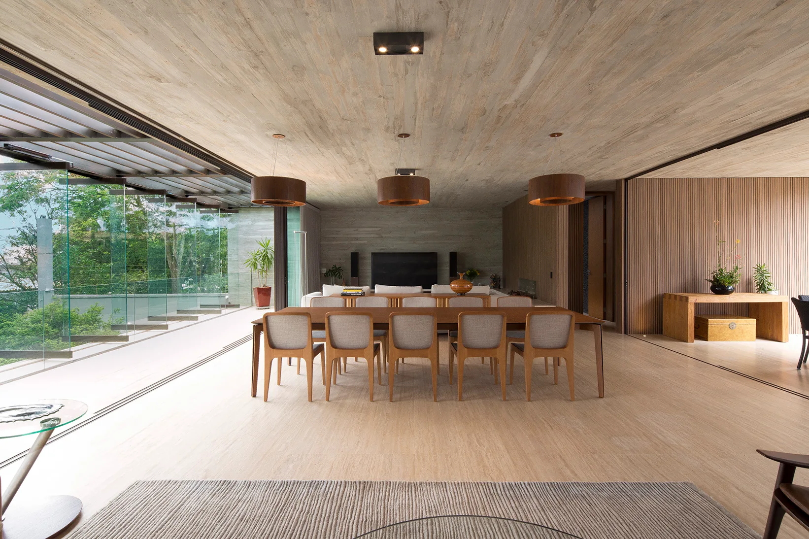 A modern dining area features a long wood table beneath three drum pendant lights, with an exposed concrete ceiling and vertical wood slat walls.