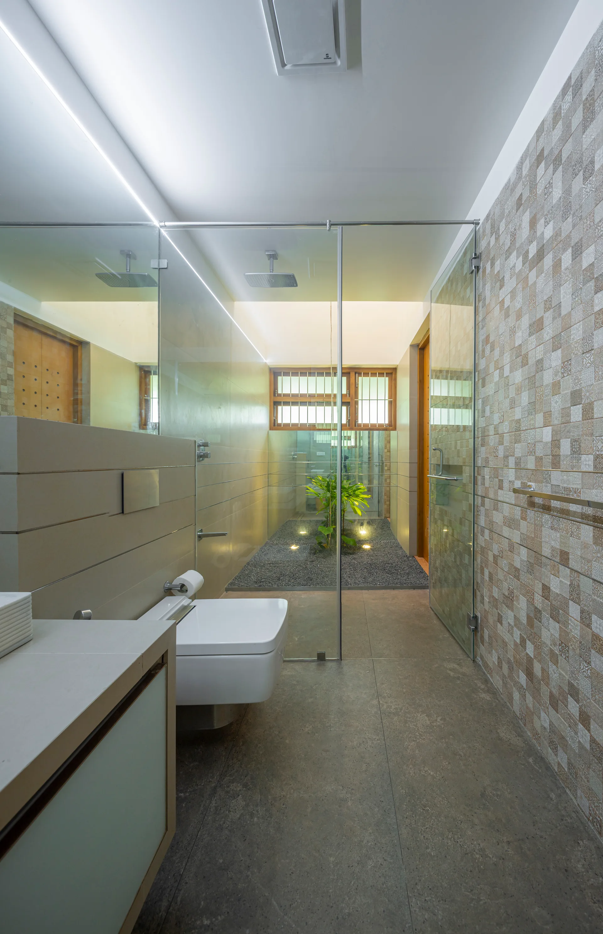A modern bathroom features a wall-mounted toilet, a glass shower partition, and a small indoor courtyard with green plants.