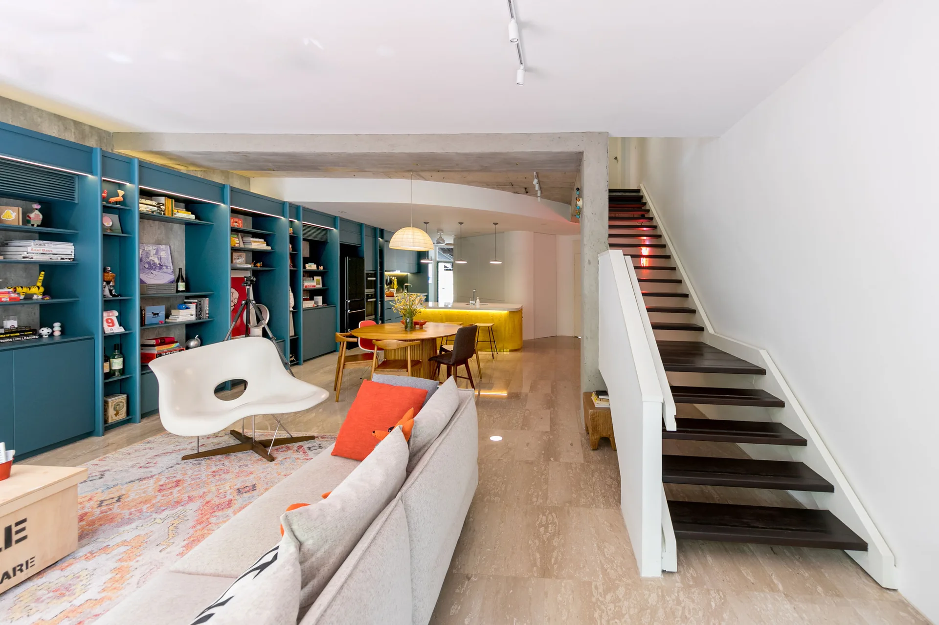A modern open-concept living room features a large teal blue storage wall, a grey sofa, a white lounge chair, and a dark wood staircase.