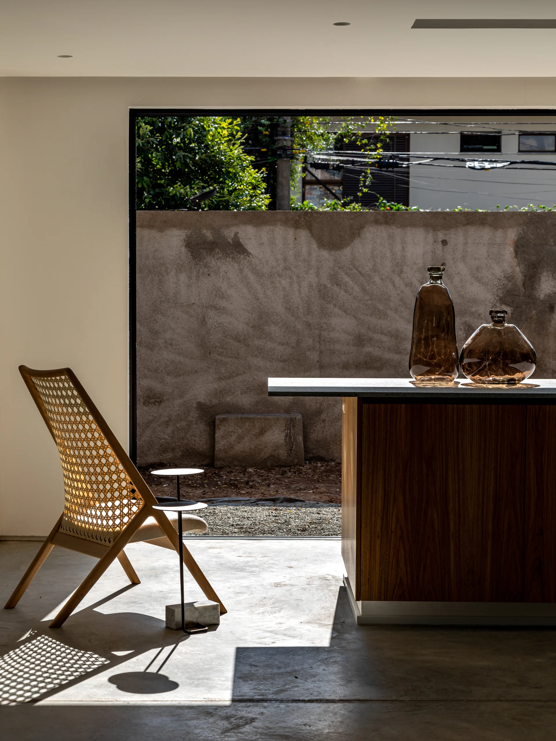 A wooden lounge chair with a woven rattan back sits on a concrete floor next to a dark wood island displaying two amber glass vases.