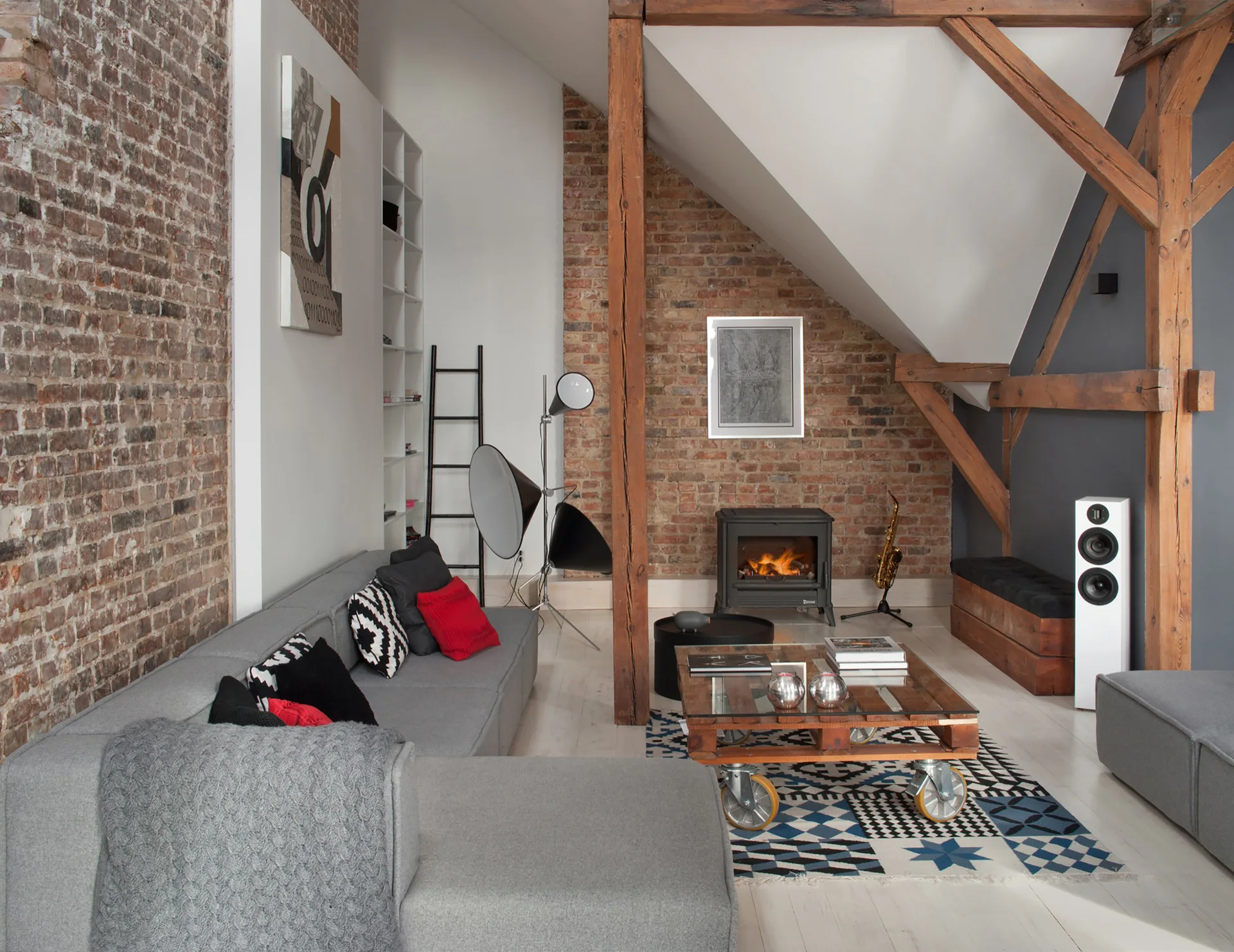 An industrial loft living room with a grey sectional, exposed brick walls, timber support beams, and a wooden pallet coffee table.