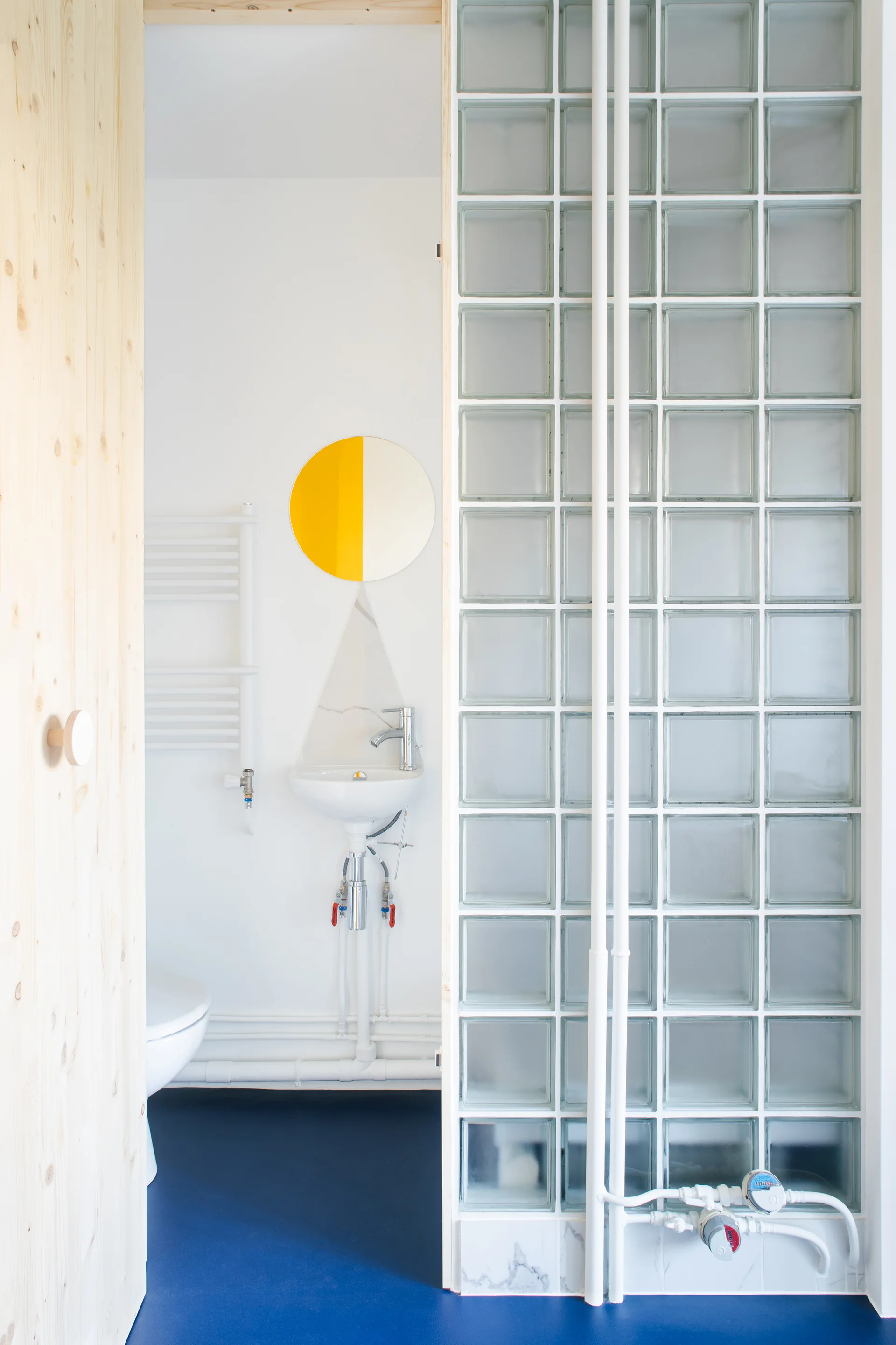 A compact white bathroom featuring a glass block partition wall, a wall-mounted sink with a yellow circular mirror, and deep blue flooring.