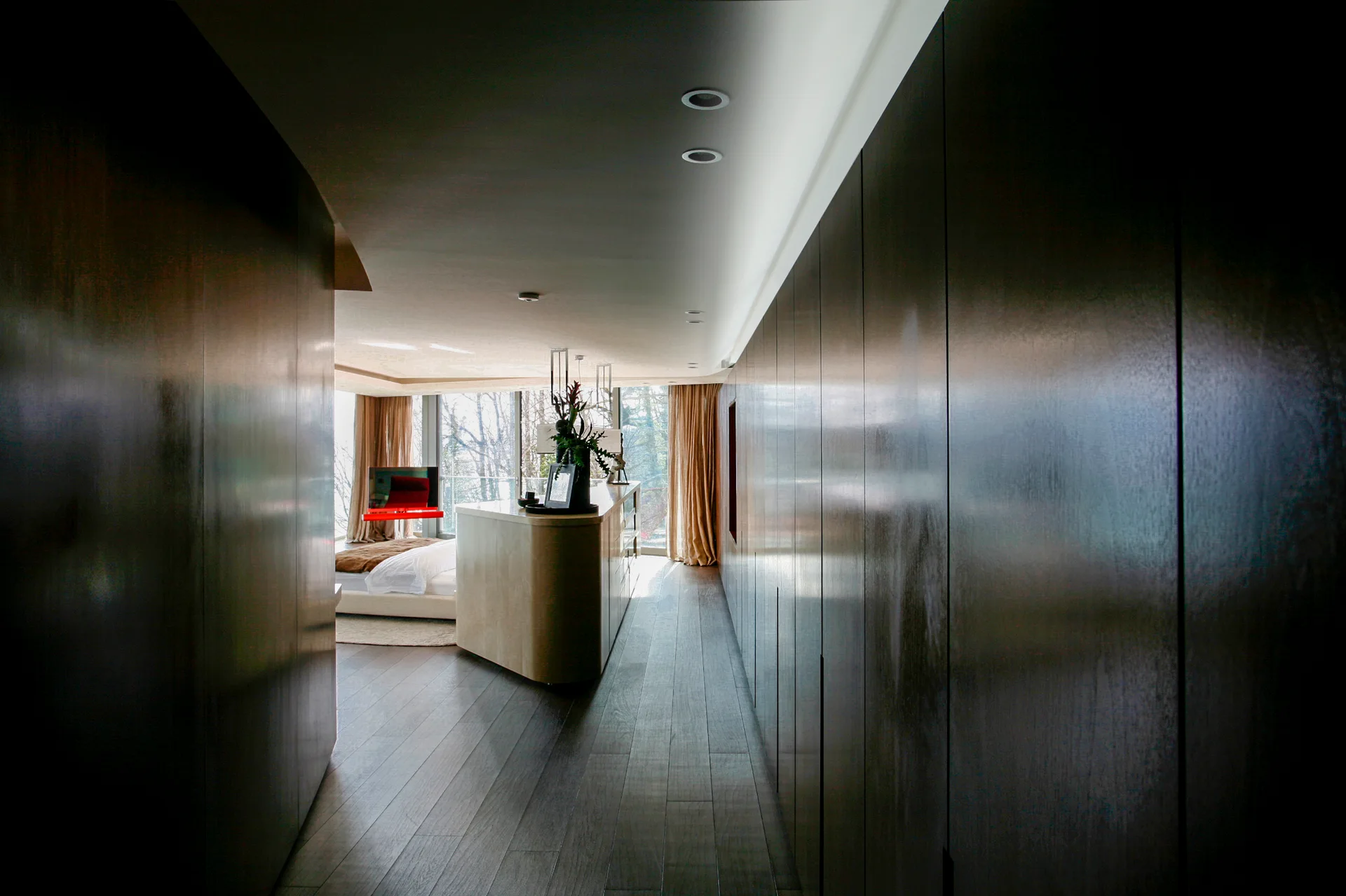 A long corridor with dark wood paneled cabinets leads to a bright bedroom featuring a curved beige console and floor-to-ceiling windows.