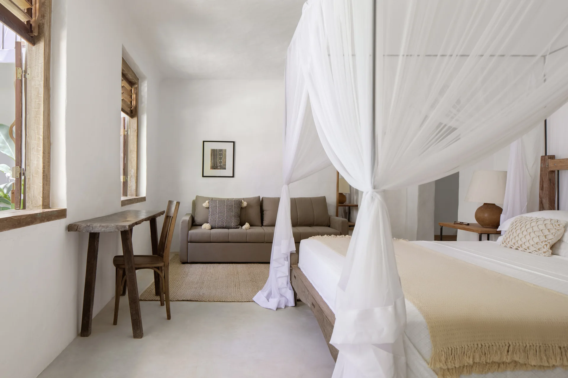 A bright bedroom featuring a canopy bed draped in sheer white netting, a rustic wooden desk by the window, and a taupe sofa in the background.