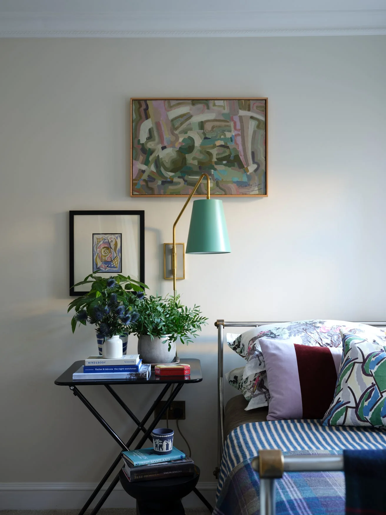 A metal bed frame with layered bedding sits next to a black tiered nightstand beneath framed artwork and a brass wall lamp with a mint green shade.