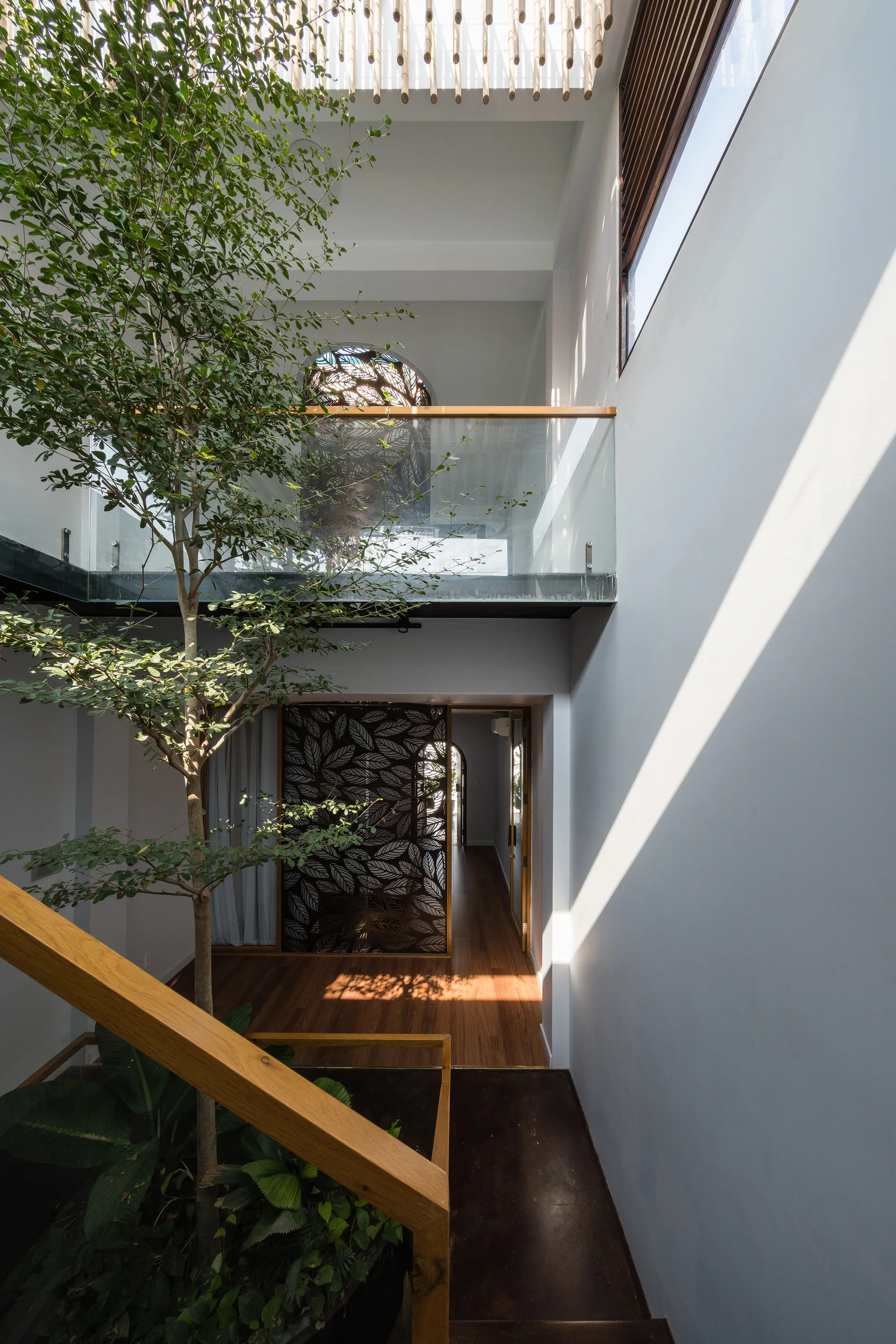 A bright multi-level atrium features an indoor tree growing past a glass-railed mezzanine, a wooden staircase, and a leaf-patterned room divider.