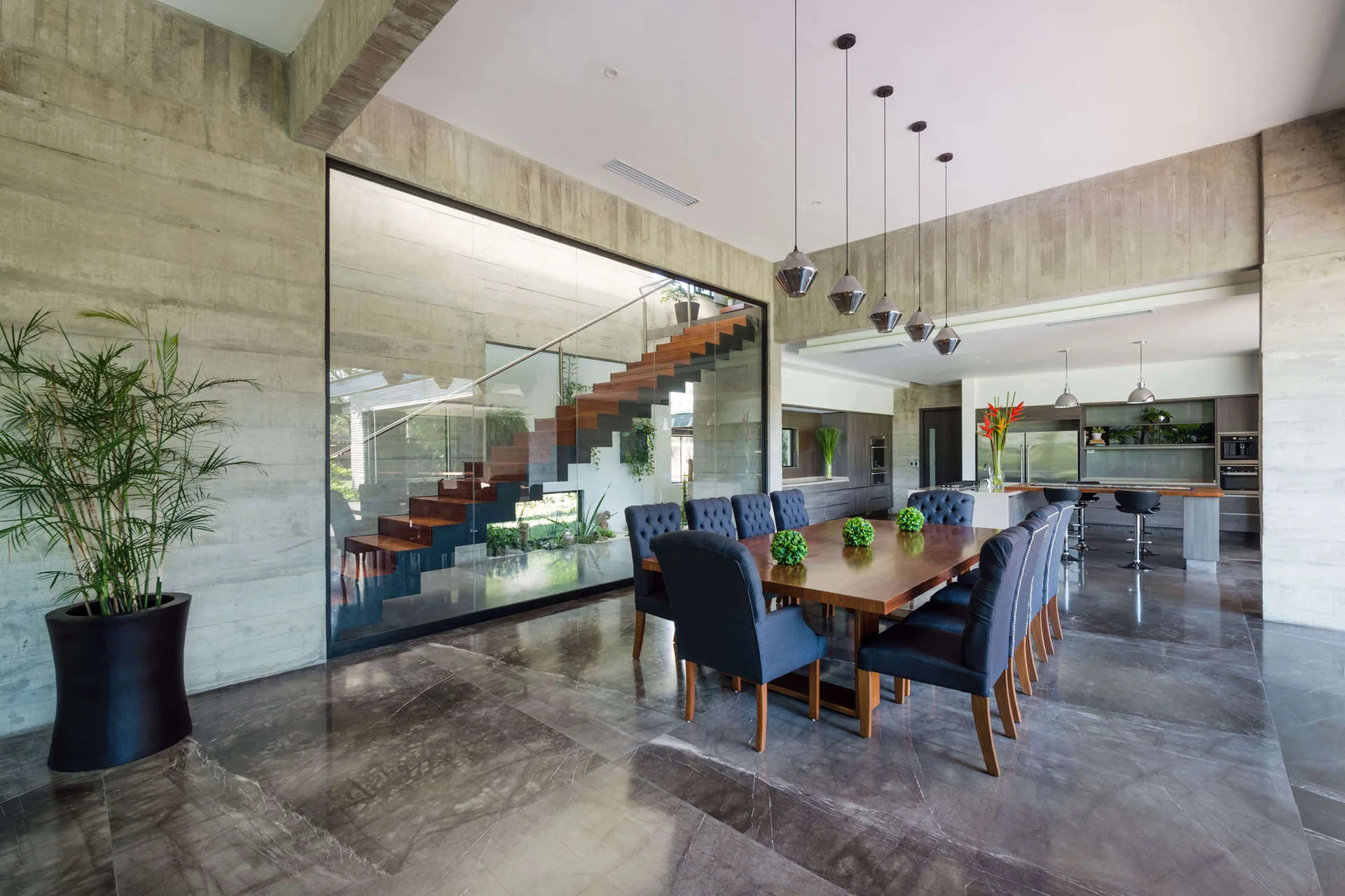 An open dining area with a wood table and dark tufted chairs set against board-marked concrete walls and a glass-enclosed staircase.