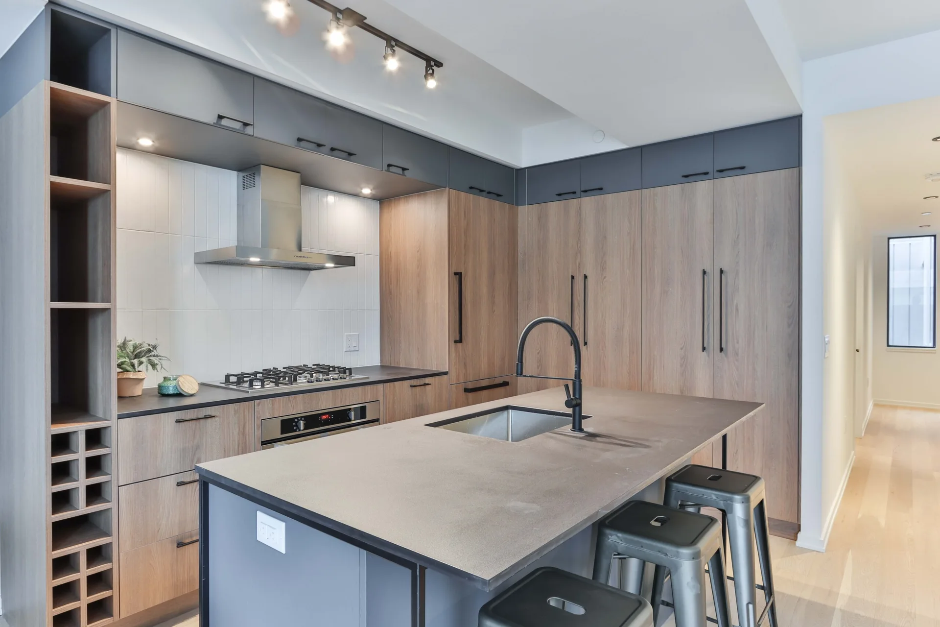 A modern kitchen features grey and wood-toned cabinets, a large island with three metal bar stools, and a vertical subway tile backsplash.