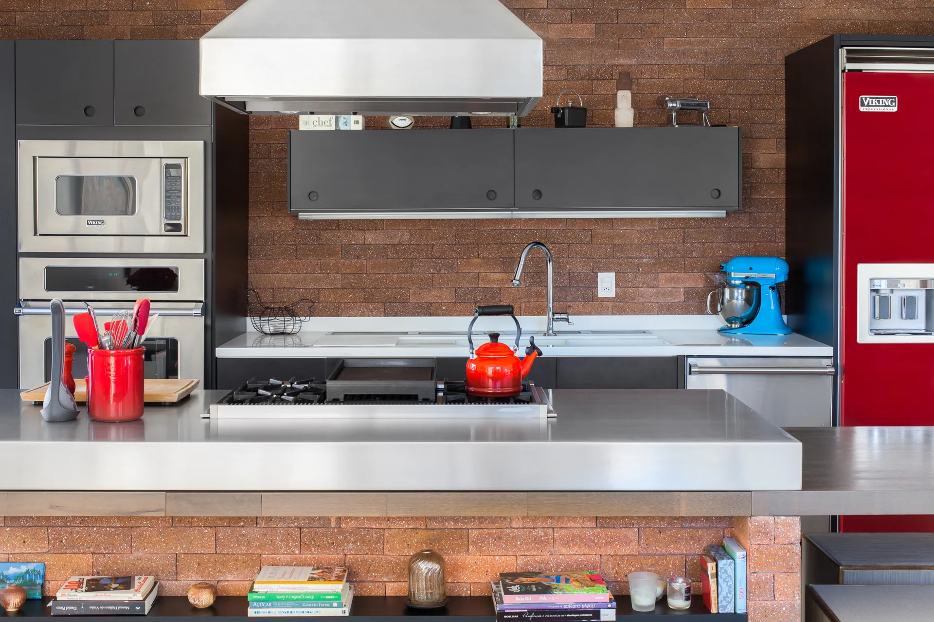 An industrial kitchen features a stainless steel island with a built-in cooktop, a red brick backsplash wall, and a bright red refrigerator.