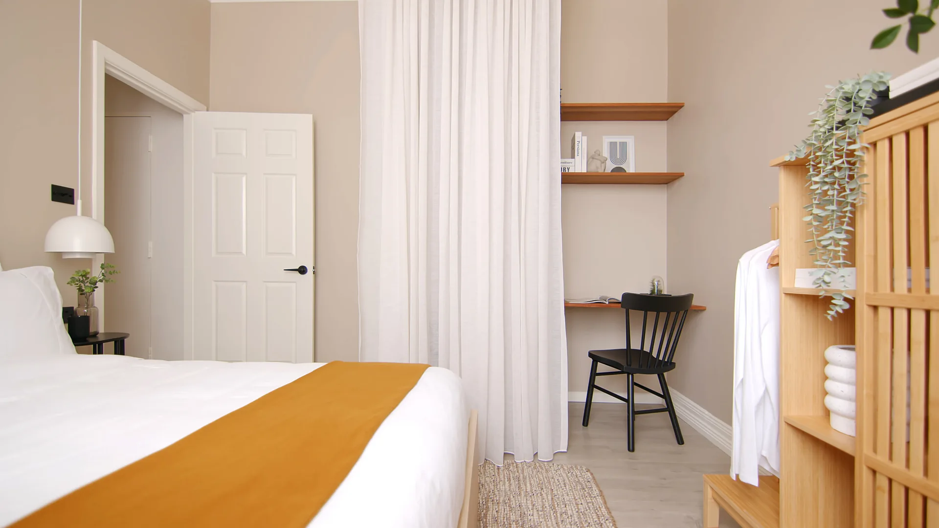 A neutral bedroom features a bed with a mustard throw, a wood closet organizer, and a small desk with a black spindle chair behind a white curtain.
