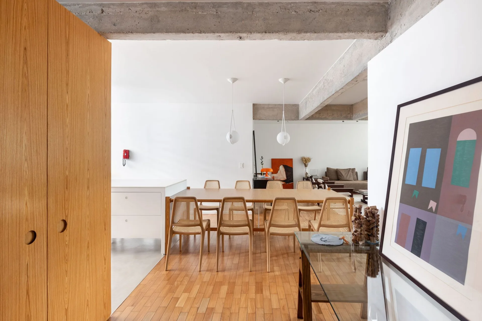 A light wood dining table with woven cane chairs stands under spherical pendant lights and an exposed concrete ceiling beam.