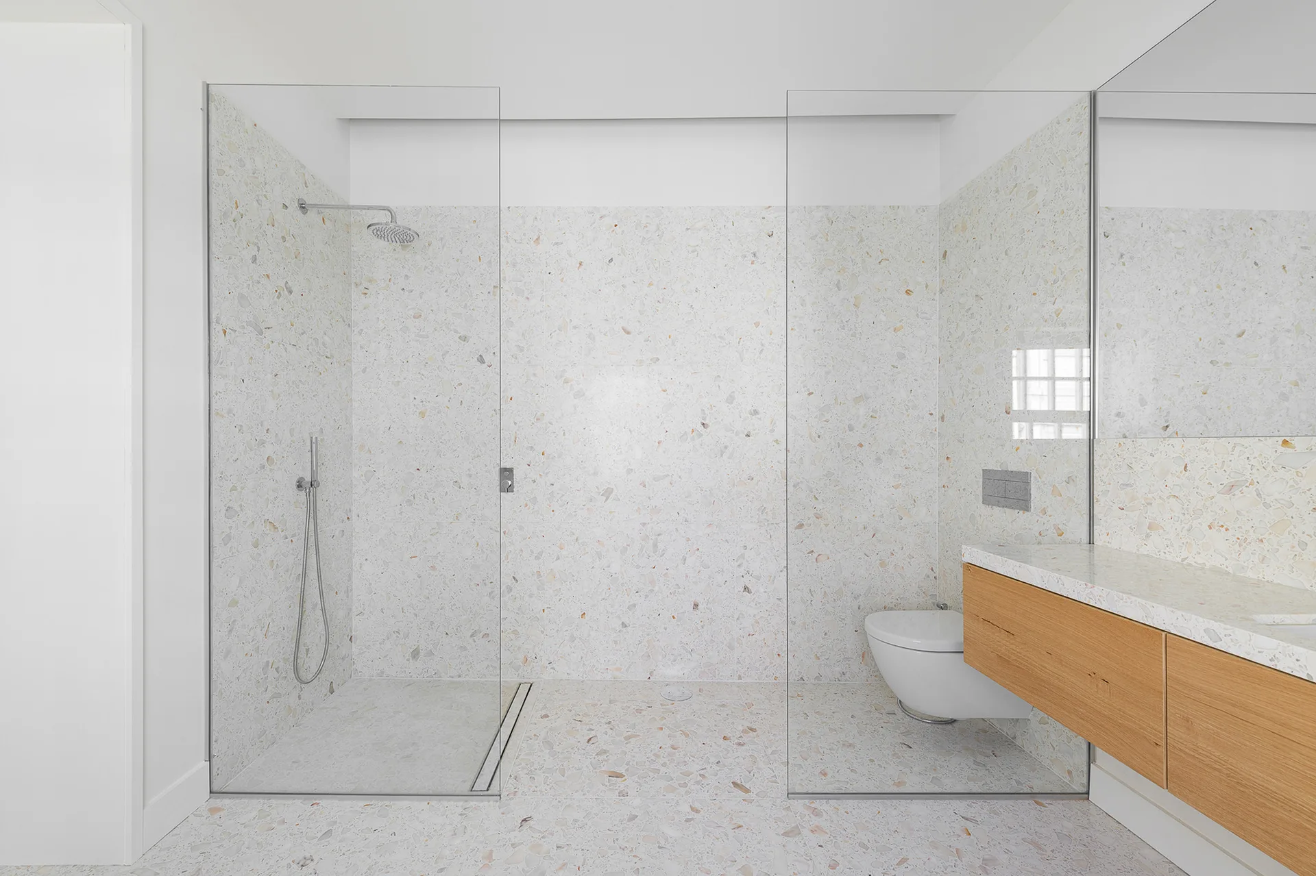 A minimalist bathroom featuring white terrazzo walls and flooring, a frameless glass shower enclosure, a wall-mounted toilet, and a light wood vanity.