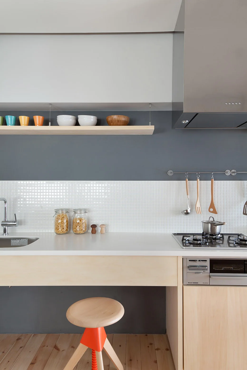 A minimalist kitchen features a white grid tile backsplash, a grey wall with a light wood floating shelf, and a wooden stool with an orange accent.