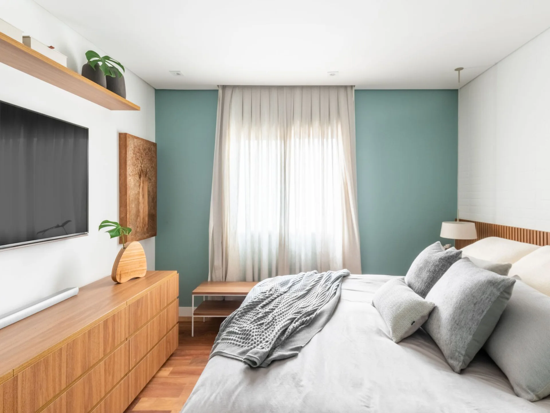 A modern bedroom features a grey bed with a slatted wood headboard next to a muted blue-green accent wall and a light wood dresser.