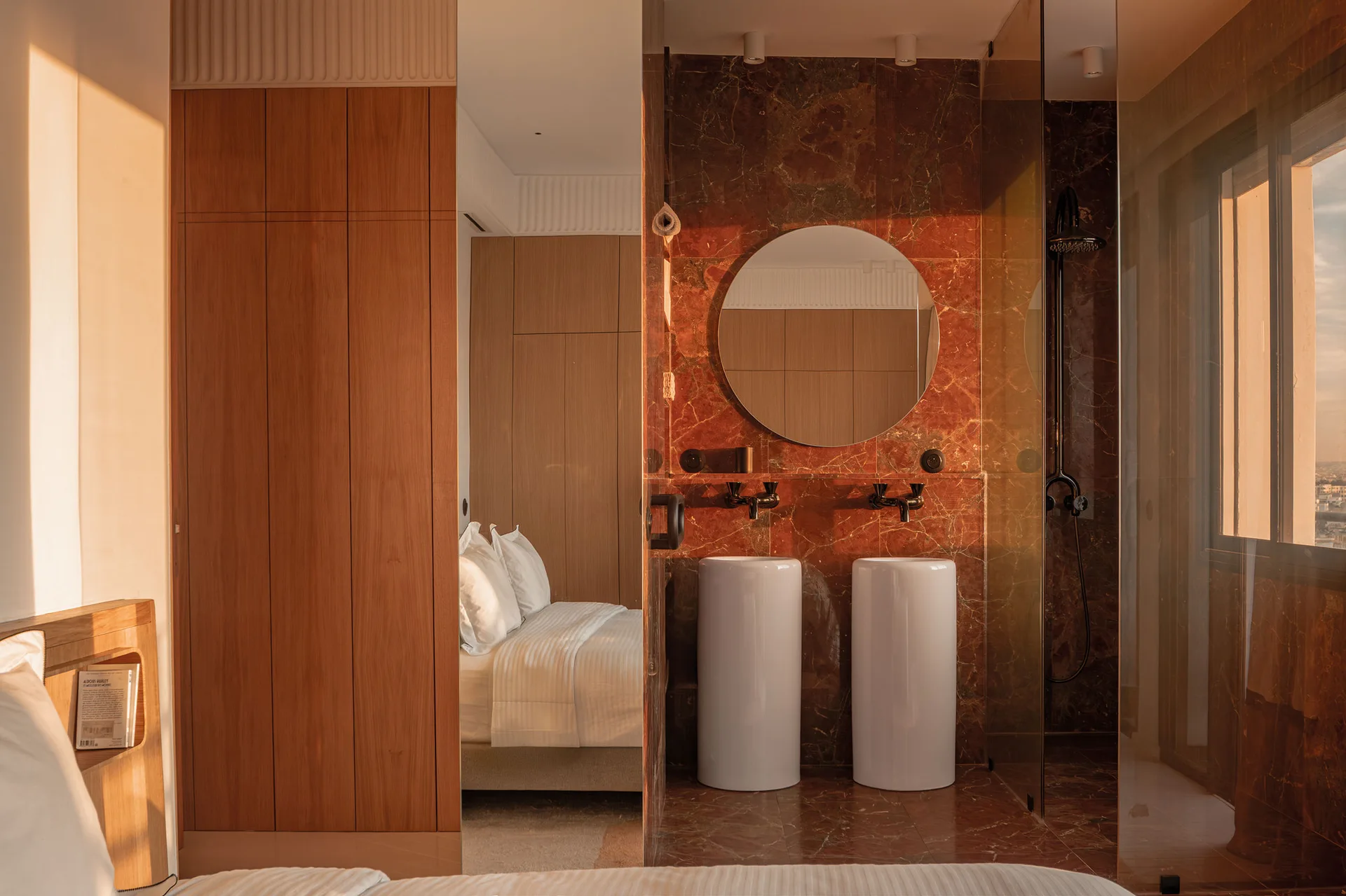 A bathroom with dark red marble walls and flooring features two white cylindrical pedestal sinks, a round mirror, and a glass partition.