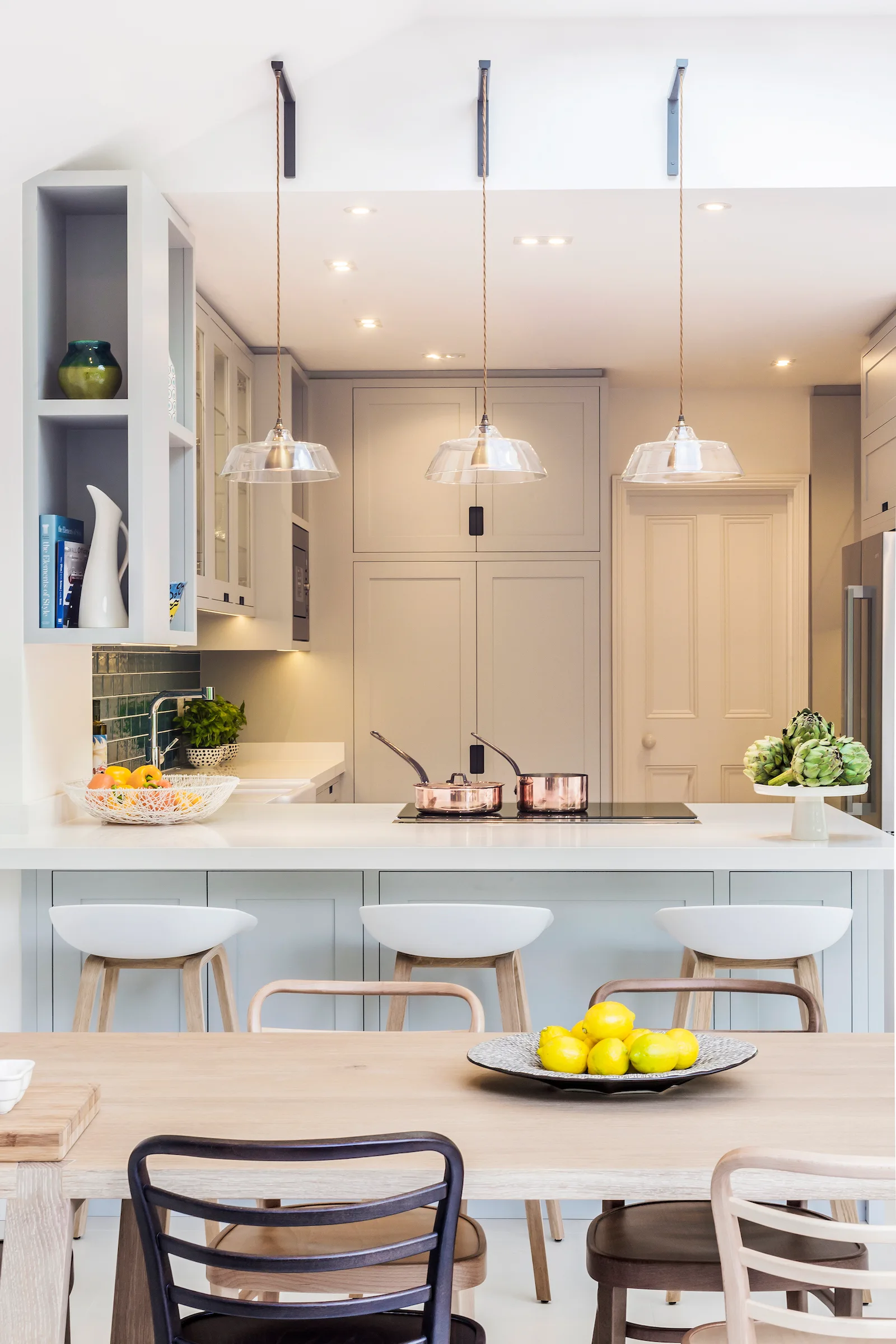 A pale grey kitchen features a central island with white bar stools, glass pendant lights, and a wooden dining table in the foreground.