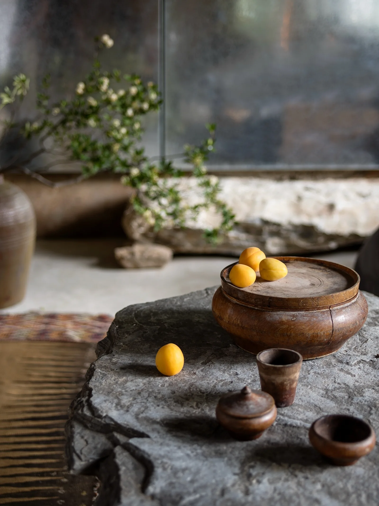 Rustic stone coffee table styled with hand-carved wooden bowls, cups, and fresh apricots in front of a textured background.