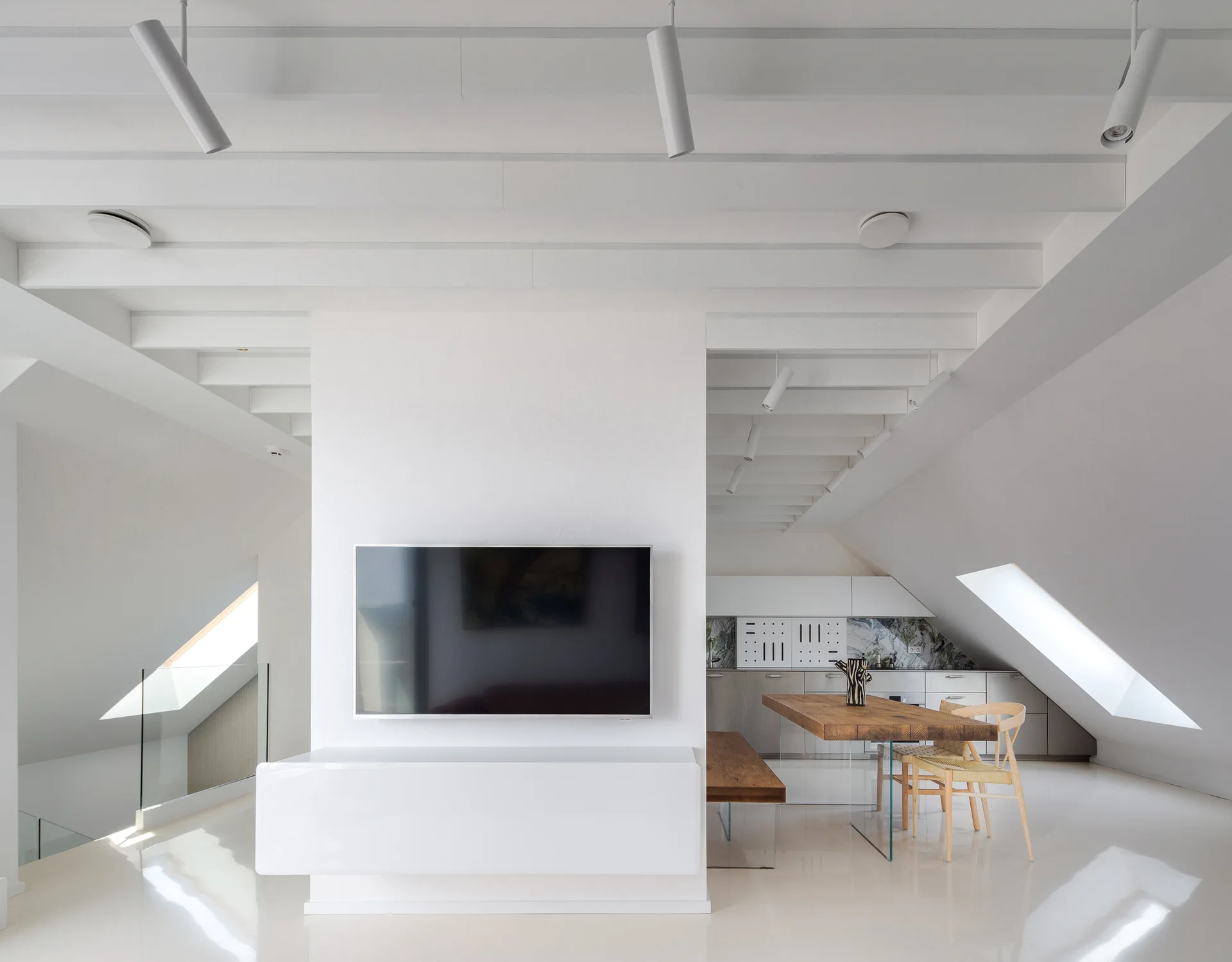 A minimalist white attic space featuring a wall-mounted television and a rustic wooden dining table with clear glass legs.