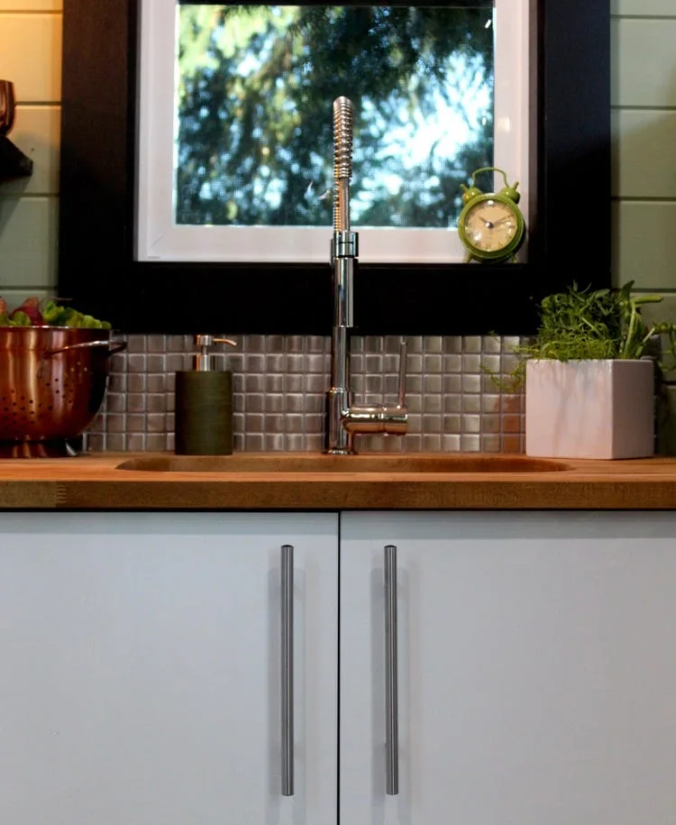 A kitchen sink area features a stainless steel faucet, wood butcher block countertop, white lower cabinets, and a reflective square tile backsplash.