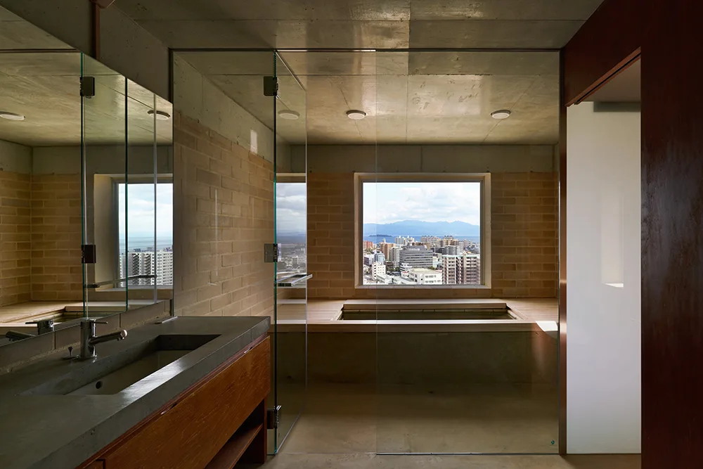 A modern bathroom features a wooden soaking tub set below a window, a concrete countertop vanity, glass partitions, and tan brick walls.