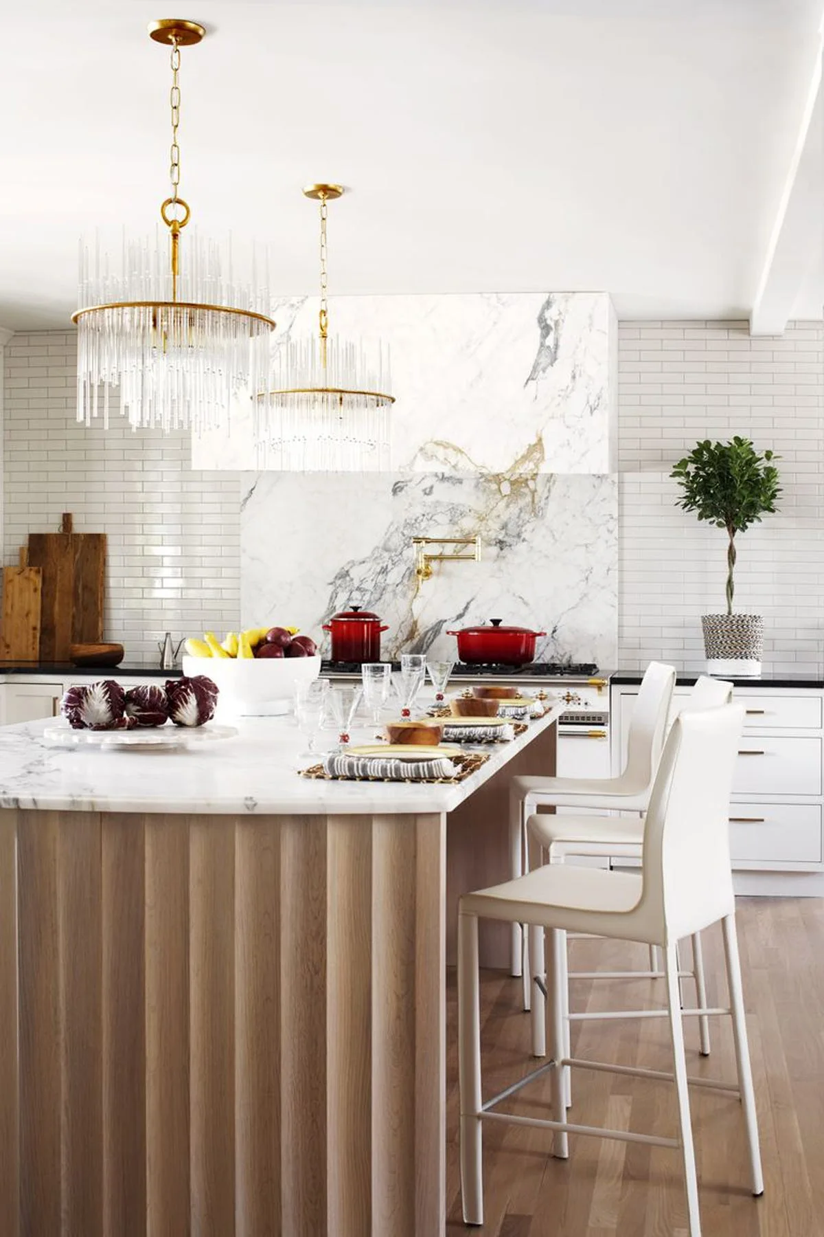 A modern kitchen features a fluted wood island with white barstools under crystal pendant lights, backed by a matching marble countertop and range hood.