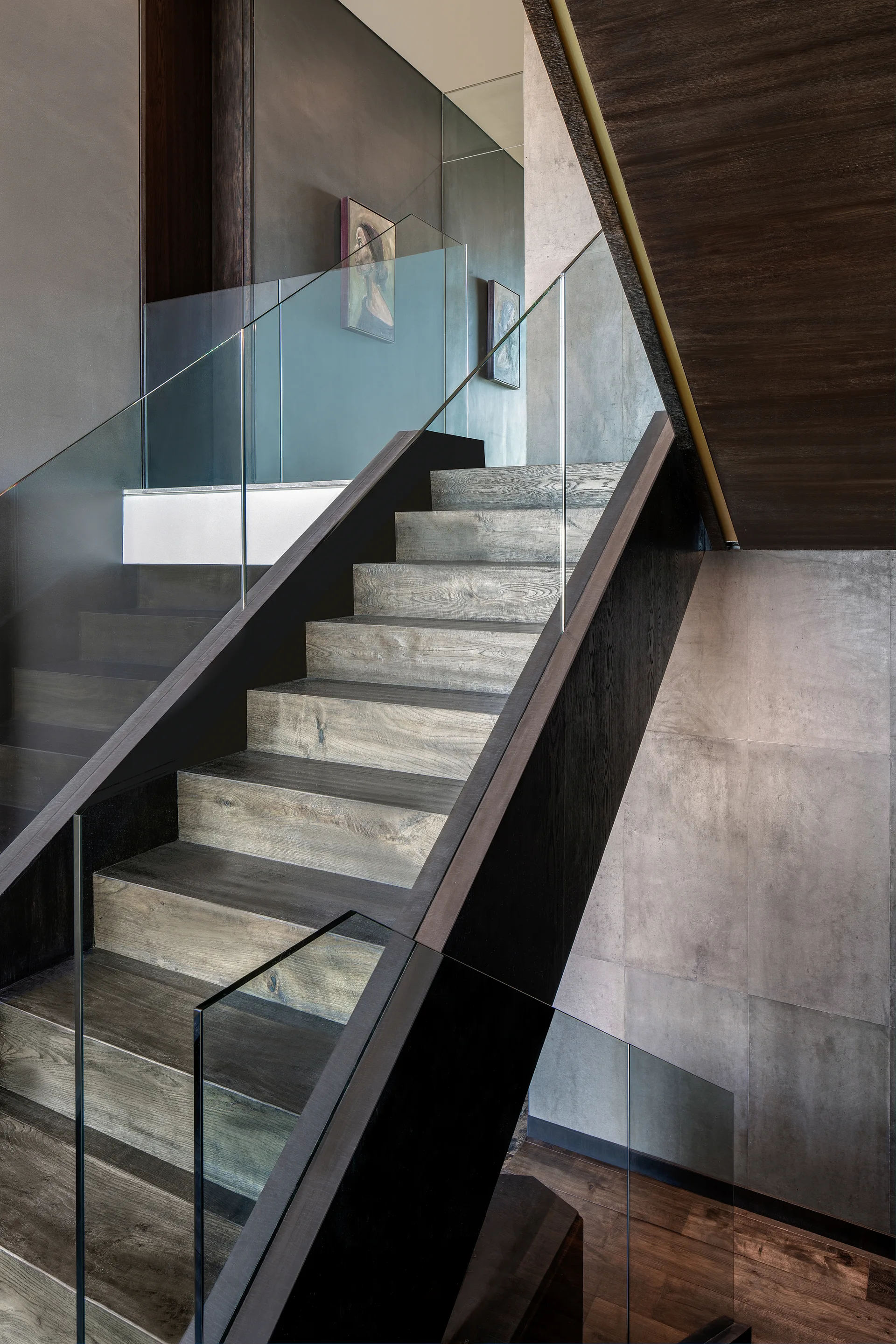 A modern staircase features textured grey wood treads, dark stringers, frameless glass railings, and concrete-look wall panels.