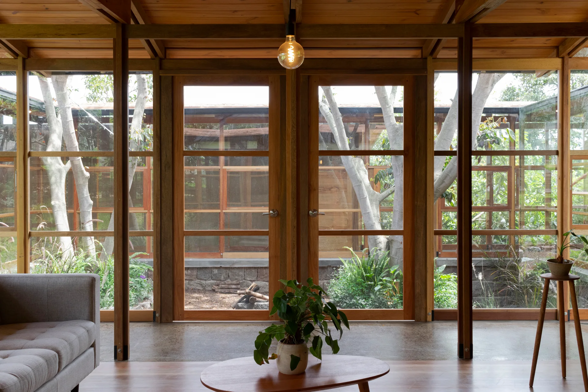 An organic modern living room features wood-framed glass double doors looking out onto a green courtyard, complete with a grey sofa and wood coffee table.